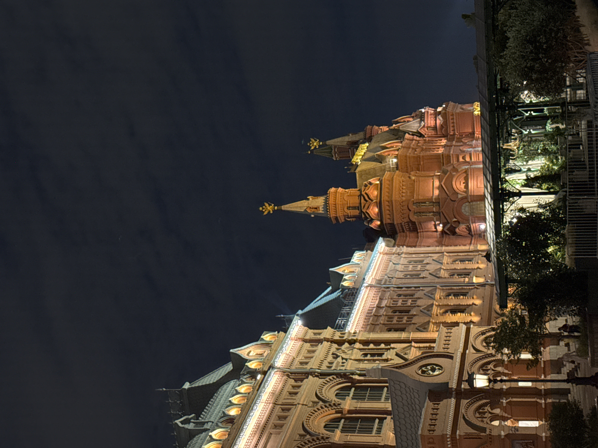 The image depicts the State Historical Museum in Moscow, Russia, at night. The museum is an imposing red brick building with intricate architectural details, illuminated by warm, artificial lights that highlight its ornate facades, arched windows, and decorative gables. Several turrets with golden finials pierce the dark night sky. In the foreground, a streetlamp casts a glow on a paved pathway, and lush green trees and foliage are visible, suggesting a park or garden area adjacent to the museum. A covered outdoor seating area with tables and chairs can be seen, hinting at a nearby restaurant or cafe. The sky is dark and cloudy, with a few faint stars visible. The overall scene conveys a sense of grandeur and historical significance, enhanced by the nighttime illumination.