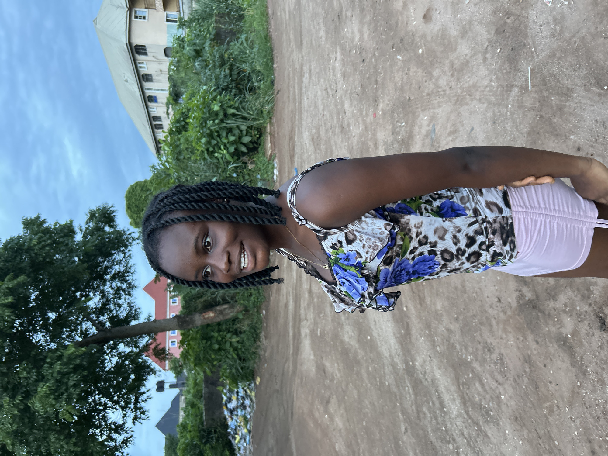 An image depicts a young Black female standing outdoors in what appears to be a residential or semi-rural area of Ihiagwa, Nigeria. The photo is taken from a slightly low angle, showing her from the waist up as she smiles broadly, looking directly at the camera. She has dark skin, brown eyes, and her hair is styled in numerous dark braids. She is wearing a sleeveless top with an animal print (leopard or cheetah) combined with striking blue floral designs. A delicate golden necklace is visible around her neck, and she appears to be wearing light pink or lavender shorts.
The background features an unpaved, dusty path or road in the midground. To the left of the path, a large tree with abundant green foliage is prominent, with parts of a red-roofed building visible behind it. Adjacent to the path on the left, a ditch contains visible scattered litter and debris. To the right of the path, dense green bushes and smaller trees line the area. Further back and to the right, a tall, light-colored building with multiple windows is visible, partially obscured by the foliage, beneath a pale blue, slightly overcast sky. The diffused light suggests either an overcast day or late afternoon. No visible text is present in the image.