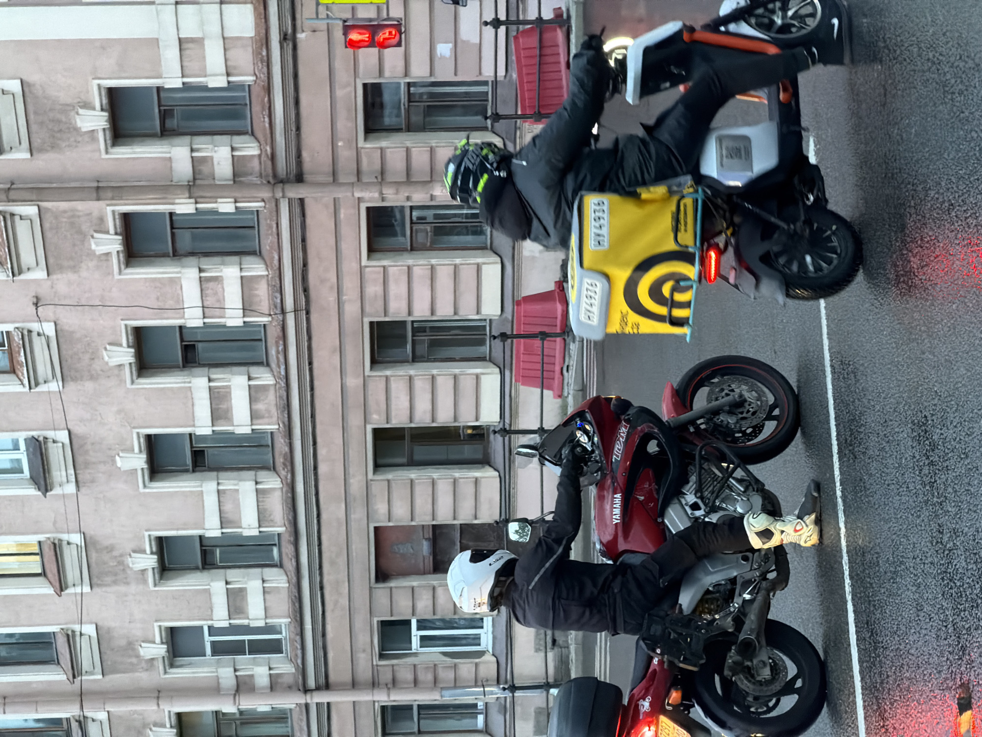 Two individuals on motorcycles are on a wet city street, likely in Saint Petersburg, Russia. The person on the left rides a red Yamaha motorcycle, wearing a white helmet and black motorcycle gear. The person on the right rides a white and orange electric scooter, with a large yellow delivery box attached to the back, and wears a black helmet. Both are moving forward on the street. In the background, a multi-story building with many windows is visible, and red traffic barriers line the sidewalk. The weather appears overcast, and the street is reflecting the lights from the vehicles and traffic signals, including a red pedestrian signal.