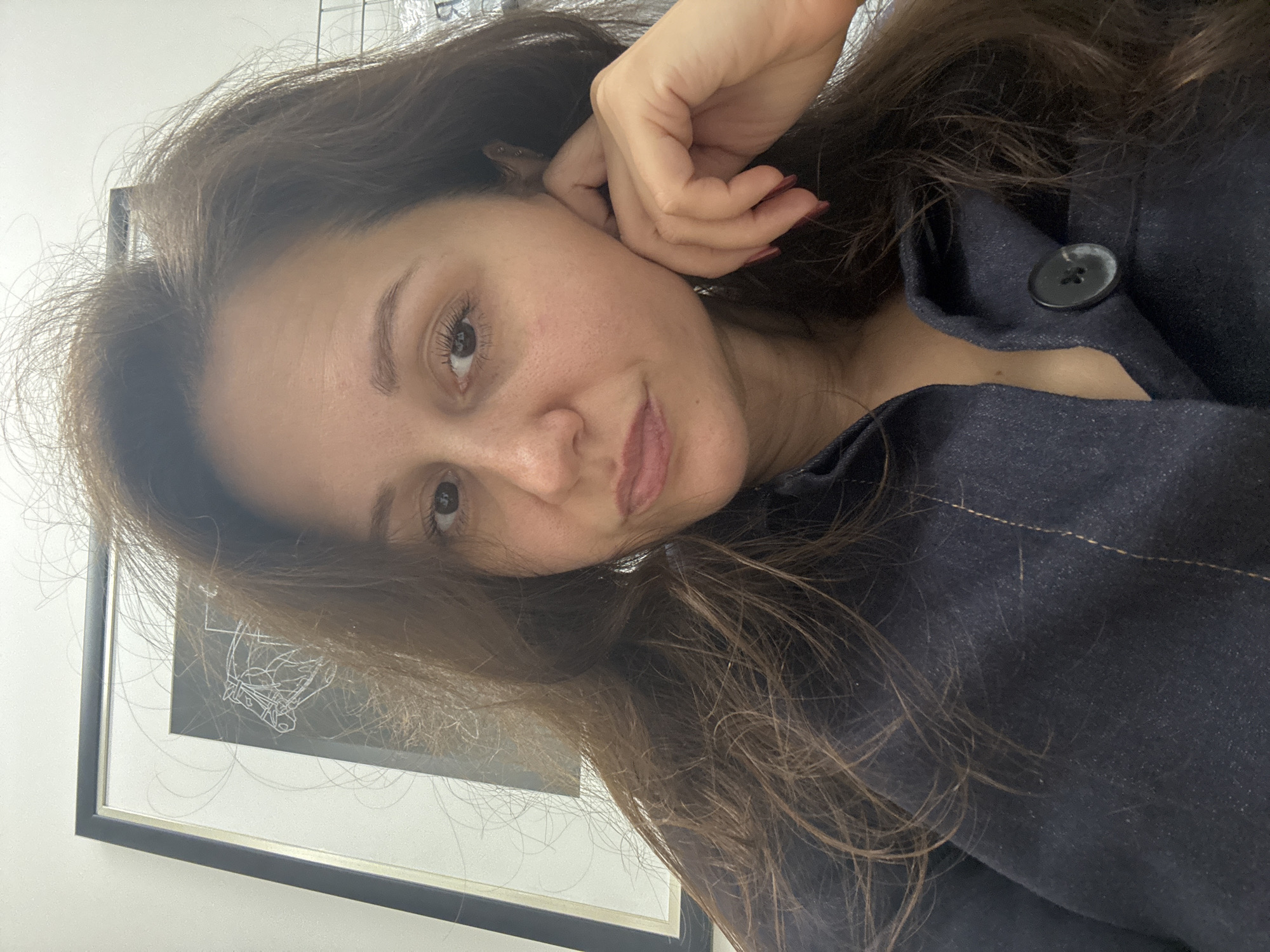 A woman with long brown hair is posing for a selfie, with her head tilted to the side and her hand near her ear. She is wearing a dark-colored collared shirt with a button visible. Behind her is a framed picture with a white sketch on a black background. The setting appears to be indoors, with a white wall in the background. There is no explicit information to suggest the photo was taken in Dubai, United Arab Emirates.