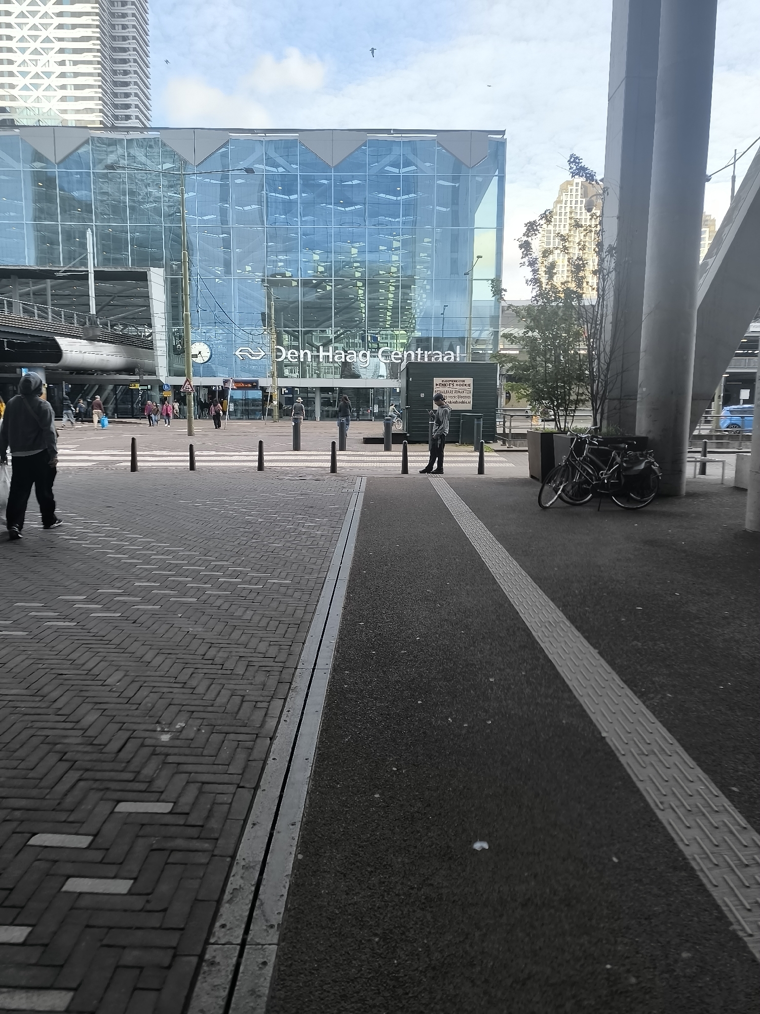 The image captures a view of Den Haag Centraal, the main railway station in The Hague, Netherlands. The modern station building, with its striking blue glass facade, is a prominent feature. In the foreground, a pedestrian area with brick paving is visible, leading towards the station entrance. A person wearing a grey hooded jacket and dark pants is walking on the left side of the pedestrian area. On the right side, a person in dark clothing is standing and looking at their phone. Two bicycles are parked near a concrete pillar on the right. The environment suggests an urban setting with a clear sky and some trees. There are also bollards lining the pedestrian crossing.