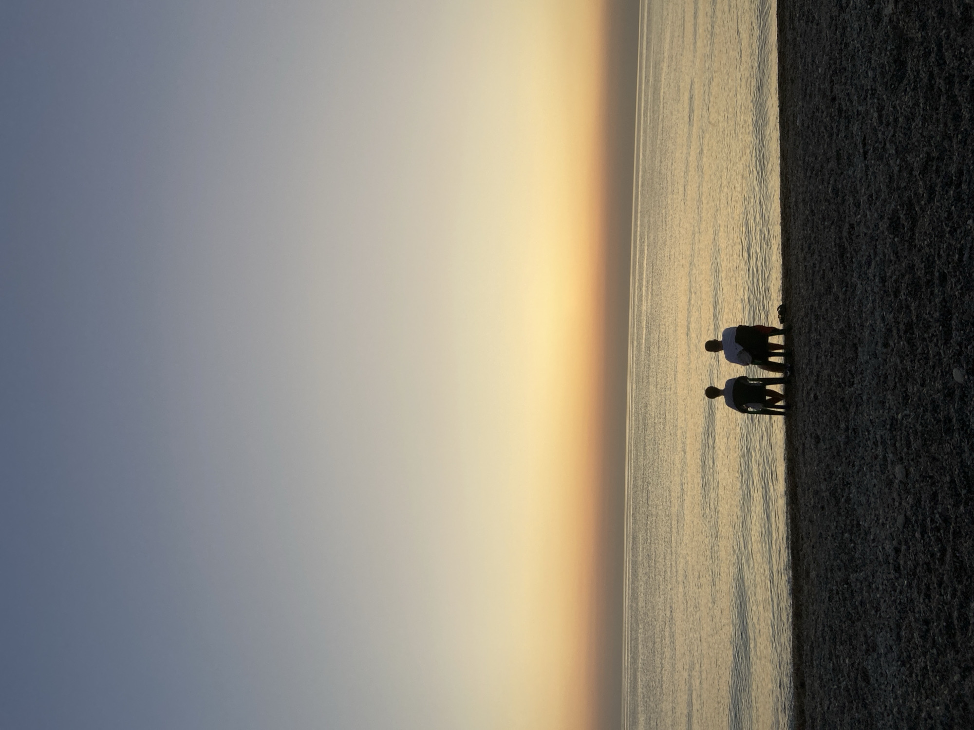 Two individuals are seated on chairs facing the sea on a pebble beach. The sky transitions from a pale blue at the top to a warm orange and yellow gradient near the horizon, indicating either sunrise or sunset. The sea is calm with gentle ripples. The scene appears to be a coastal area in Sevastopol, Ukraine, given the typical landscape and the calm sea conditions often observed there. The individuals are silhouetted against the bright sky and sea, obscuring their facial features and detailed attire. They are positioned side-by-side, suggesting a shared experience of observing the seascape. The overall mood is tranquil and contemplative.