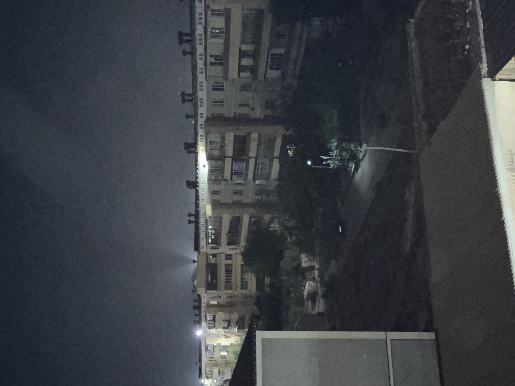 The image captures a nighttime view of an urban residential area, likely in Fergana City, Uzbekistan, as suggested by the provided context. The scene is dominated by multi-story apartment buildings, characterized by their drab, light-colored facades and numerous balconies. The architecture suggests a Soviet-era design, common in many cities within Central Asia. The sky is dark and overcast, with the only illumination coming from streetlights and lights emanating from within the apartment windows.
In the foreground, a dark, sloped roof of a lower building is visible, with a lighter-colored, multi-story structure partially obscuring the view to the left. Beyond these, the apartment buildings stretch horizontally across the frame. Trees and vegetation are interspersed between the buildings and in front of them, their forms softened by the dim lighting and creating shadows. A lone streetlight stands on the right side of the frame, casting a pool of light on the ground and illuminating a small, bushy plant. The overall impression is one of a quiet, dimly lit urban environment at night. No people are visible in the frame.