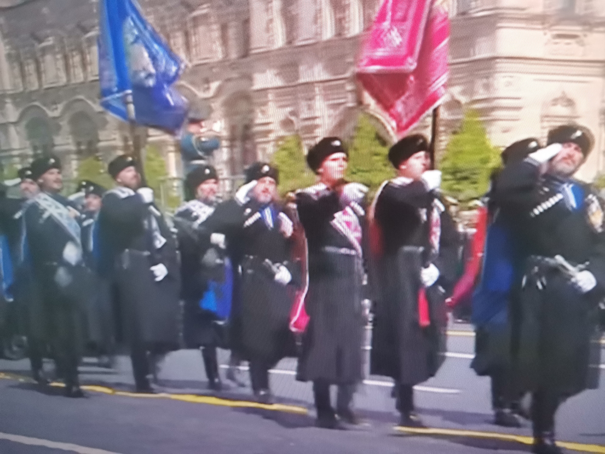 The image shows a parade with several people in uniform. They are wearing dark coats and hats, and some are carrying flags. The flags are large and colorful, one is blue and the other is pink. The people appear to be marching in formation on a paved street. In the background, there are buildings with arches. The lighting suggests it is daytime. The scene is consistent with a formal event or ceremony.
