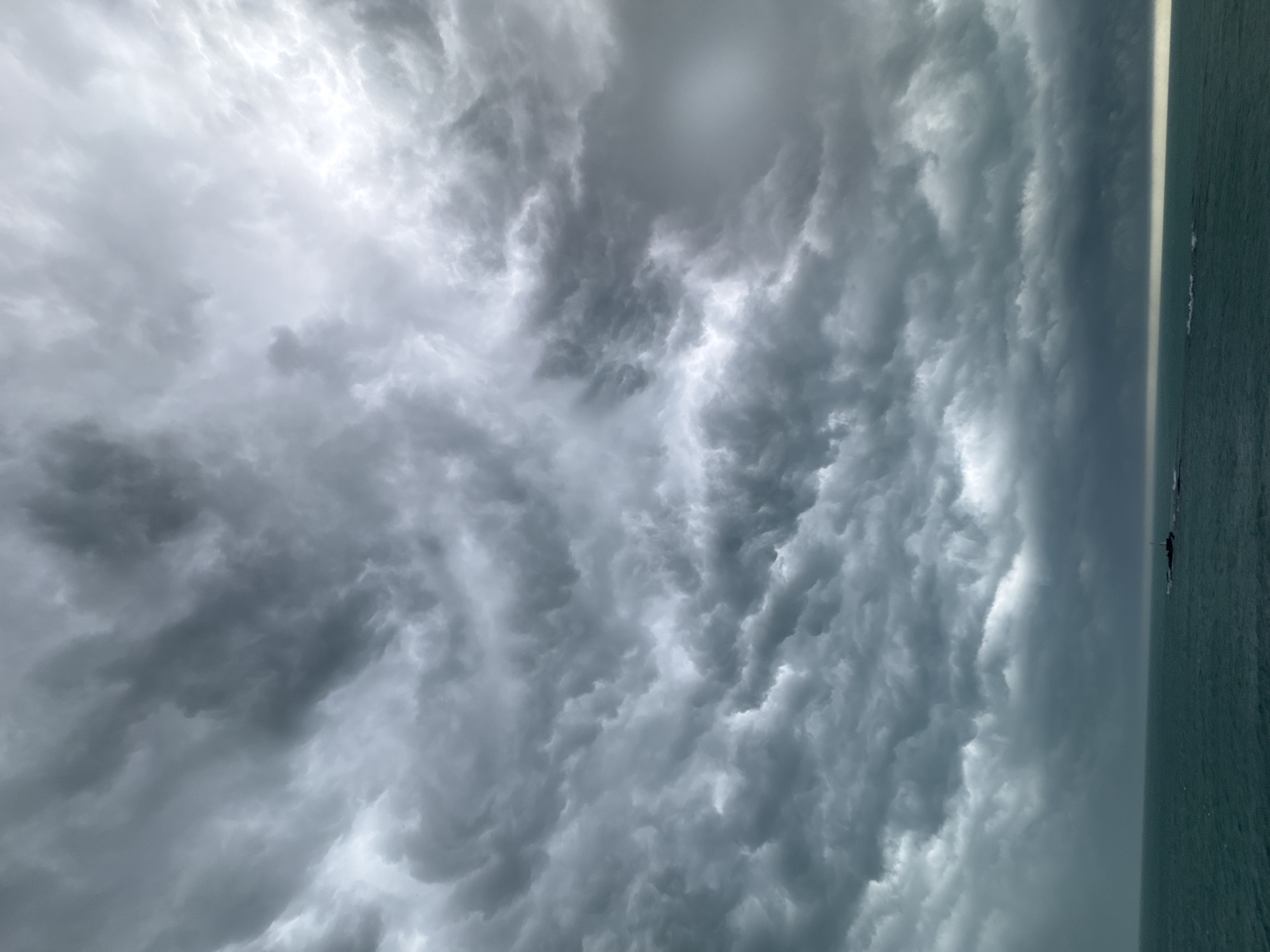 This photograph presents a dramatic coastal scene, likely captured in Tsarevo, Bulgaria, overlooking the Black Sea. The image is dominated by an expansive sky filled with powerful, turbulent storm clouds, occupying approximately two-thirds of the frame. These clouds vary in shade from deep charcoal to lighter greys, with bright patches where sunlight attempts to pierce through, highlighting their intense, undulating textures. The formations appear heavy and ominous, suggesting an active or impending weather event, possibly a thunderstorm or a heavy overcast condition.
A striking, intensely bright horizontal line cuts across the image, sharply separating the turbulent cloudscape from the dark expanse of the sea below. This horizon line creates a stark contrast, almost glowing against the muted tones of the sky and water. The lower third of the image features the sea, appearing a deep greenish-blue, reflecting the cloudy conditions above. The water's surface shows subtle variations, hinting at gentle waves.
Along the bright horizon line, two very small, dark silhouettes are visible. One clearly depicts a person standing on what appears to be a paddleboard, while the other is a less defined dark shape, possibly another distant individual or object. Their tiny scale emphasizes the vastness and power of the surrounding natural elements. The scene conveys a sense of raw natural beauty and dramatic weather, likely captured during an overcast day or during a specific time when the sun's position creates such a luminous horizon effect against the dark clouds. No visible text is present in the image.