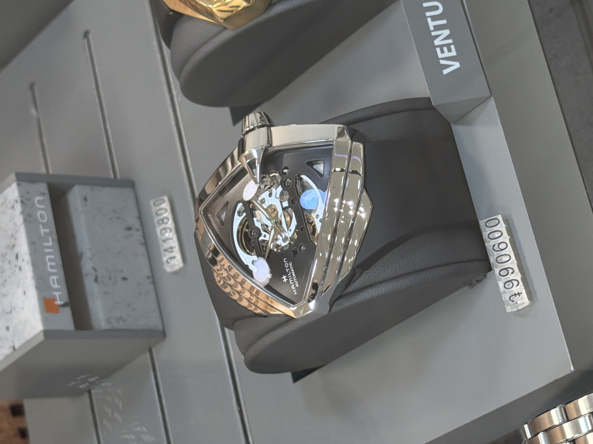 A close-up shot shows a Hamilton watch displayed in what appears to be a retail setting. The watch has a unique, shield-shaped design with a visible automatic movement. The model is named "Ventura". The watch is mounted on a gray display stand with a price tag of "€990600". Another tag with the number "3419800" is partially visible on the left. The location is Yerevan, Armenia.