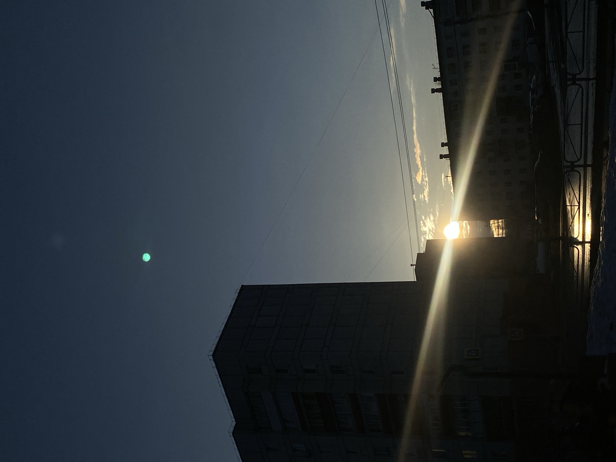 This image appears to be a low-angle shot taken in Chaikovsky, Russia, depicting a skyline dominated by apartment buildings. The sun is positioned low on the horizon, creating a bright glare and casting long shadows. The sky is a mix of dark blue and lighter hues near the horizon. Overhead wires are visible against the sky. A noticeable green orb is present in the upper-left portion of the image, possibly a lens flare. The buildings are silhouetted against the bright sunlight, revealing their blocky shapes and regular window patterns. Some snow is visible in the bottom-right corner. The image is oriented vertically.