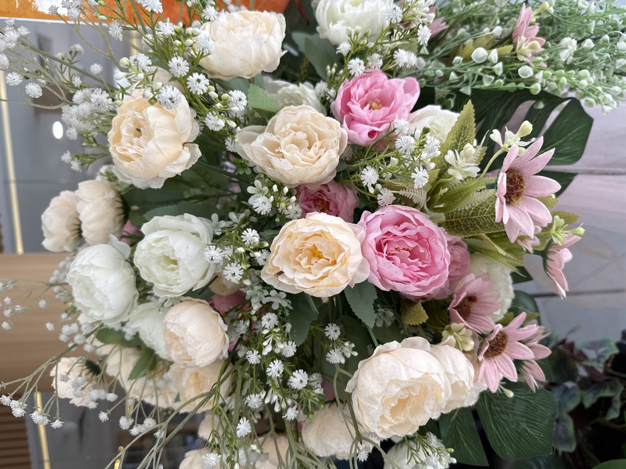A close-up, vertical shot showcases a vibrant floral arrangement, likely artificial, positioned indoors. The bouquet is a medley of peony-style flowers in soft shades of cream, peach, and pink, interspersed with delicate white baby's breath and other small, white blossoms. Green foliage, including large monstera leaves, provides a lush backdrop. The arrangement is densely packed, suggesting it's a centerpiece or decorative display. The lighting is bright, illuminating the textures and colors of the flowers. The background is blurred but hints at a modern interior with wooden elements and possibly glass. The specific location in Kuta, Indonesia, is not discernible from the visual information provided in the image.