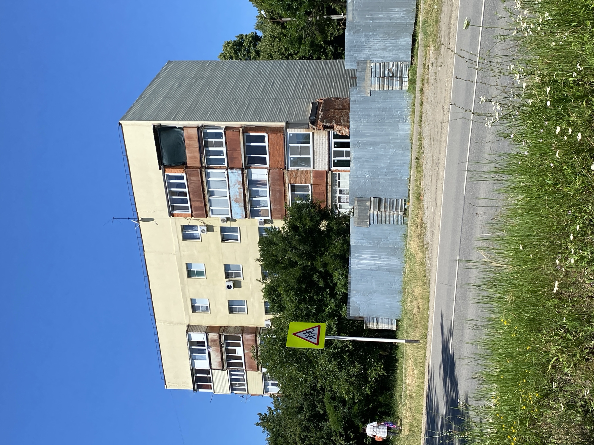 The image depicts a residential building in the city of Zapadny, Russia. The building is a multi-story apartment complex with a light yellow facade and red brick accents around some of the windows and balconies. The building appears to be old, with some signs of wear and tear, including rust on the balconies and exposed brick on one corner. A large metal fence runs along the front of the property, separating it from a paved road. A pedestrian crossing sign with a silhouette of children is visible on a pole near the road. A woman is walking away from the camera on the grassy area in front of the building, carrying a bag. The sky is clear and blue, suggesting a sunny day. Lush green trees surround the building.
