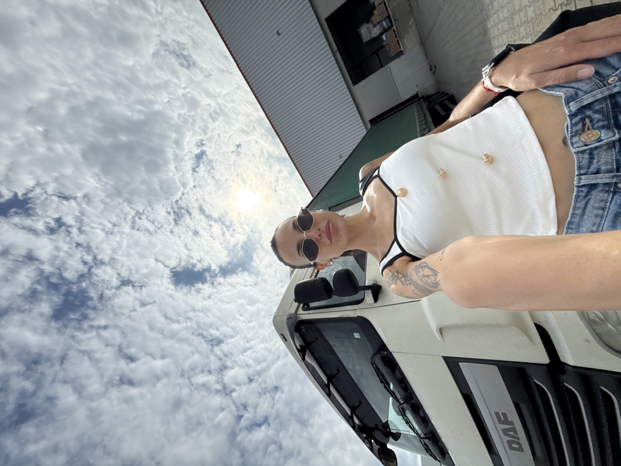 A woman with sunglasses stands in front of a white DAF truck. She is wearing a white ribbed tank top, denim shorts, and a watch on her left wrist. She has tattoos on her left arm and is wearing a necklace. The truck is a DAF model, and the grill is visible. In the background, there is a building with a corrugated metal roof. The sky is cloudy. The image was taken in Pleven, Bulgaria.