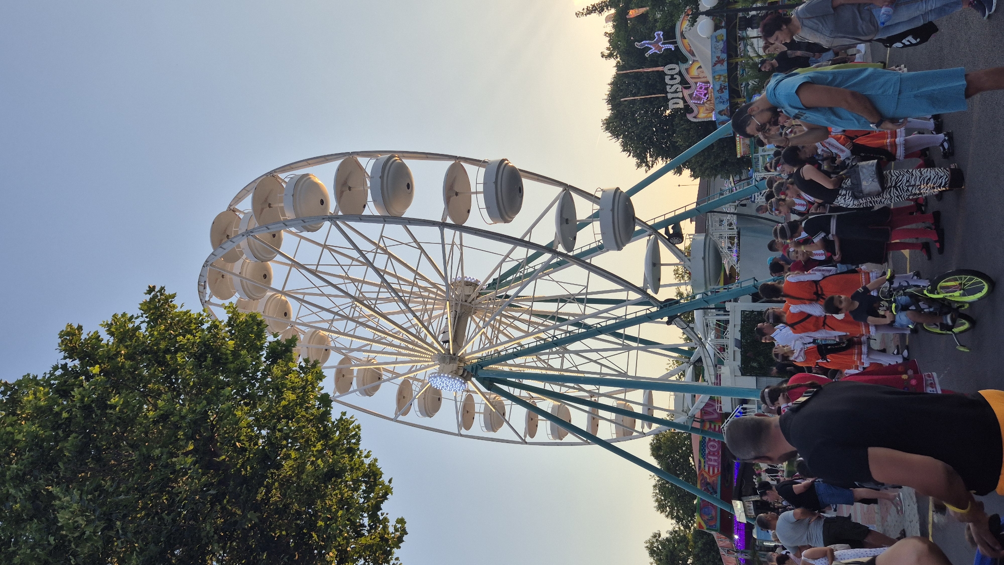 This is a medium shot, taken outdoors at dusk, of a large Ferris wheel in an amusement park. The Ferris wheel is white with teal accents and has numerous white gondolas. A crowd of people, some in traditional Bulgarian folk costumes, are gathered around the base of the Ferris wheel, and to the right of the image is a "DISCO" sign with flashing lights. The sky is a clear, pale blue, and the setting sun is visible on the right side of the image, casting rays of light. The general atmosphere is festive and lively. The location is Pomorie, Bulgaria.