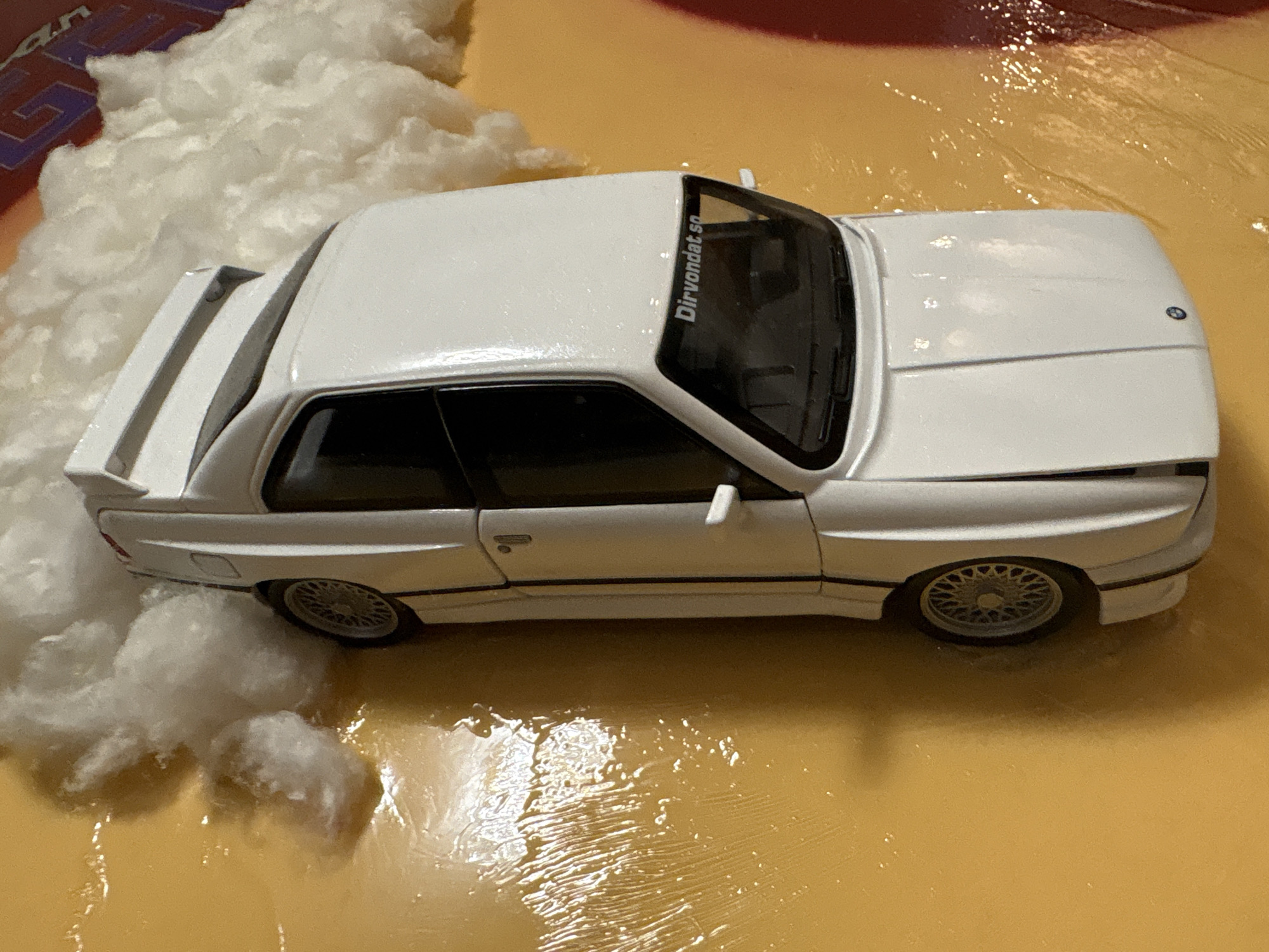 A white toy car is positioned on a yellow surface that appears to be wet or has a glossy finish. The car is facing downwards and slightly to the left. It has black windows and a spoiler on the back. There are fluffy white objects, resembling cotton or clouds, scattered around the car and on the yellow surface. The car has the text "Dirvondat.se" written on its rear window. The background includes a reddish-brown colored object with some text visible that appears to be part of a logo. The scene is lit with artificial light, casting some reflections on the wet surface.