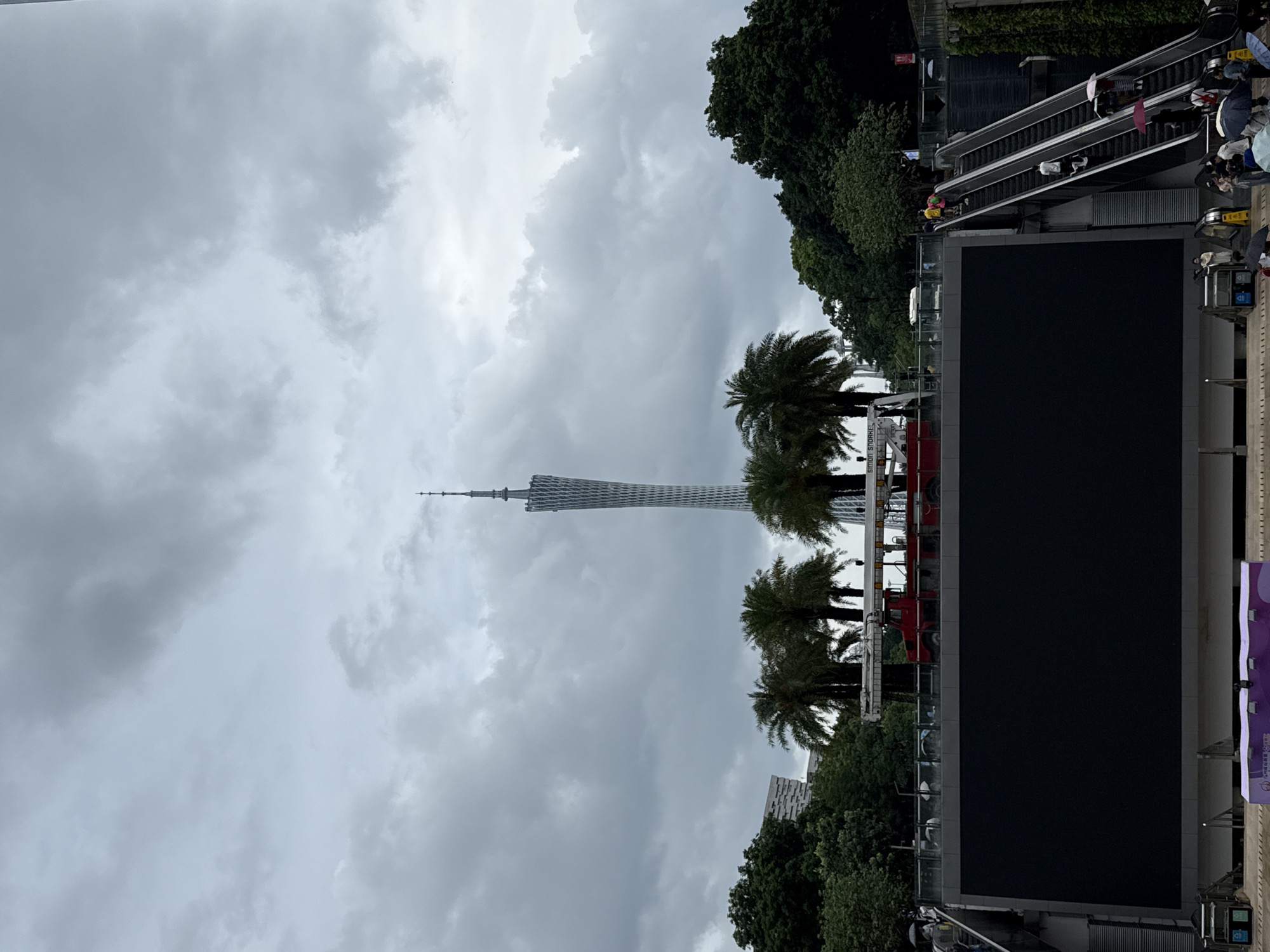 This image captures a wide-angle view of an outdoor urban scene with a prominent tower in the background under a cloudy sky. The tower, identifiable as the Canton Tower in Guangzhou's Tianhe District, China, is a slender, latticed structure that tapers towards its apex. In the middle ground, a large, dark, rectangular screen dominates the foreground, possibly a digital billboard or display. Above the screen, a red fire truck with an extended ladder is visible, suggesting maintenance or a staged event. Several palm trees and dense green foliage frame the scene, with buildings of varying architectural styles peeking through the trees. People are dispersed throughout the image, with some ascending an escalator on the right side, holding umbrellas, indicating potential light rain. Others are seen near the screen and in the background, contributing to the sense of a public space. The overall atmosphere is overcast, contributing to a muted color palette.