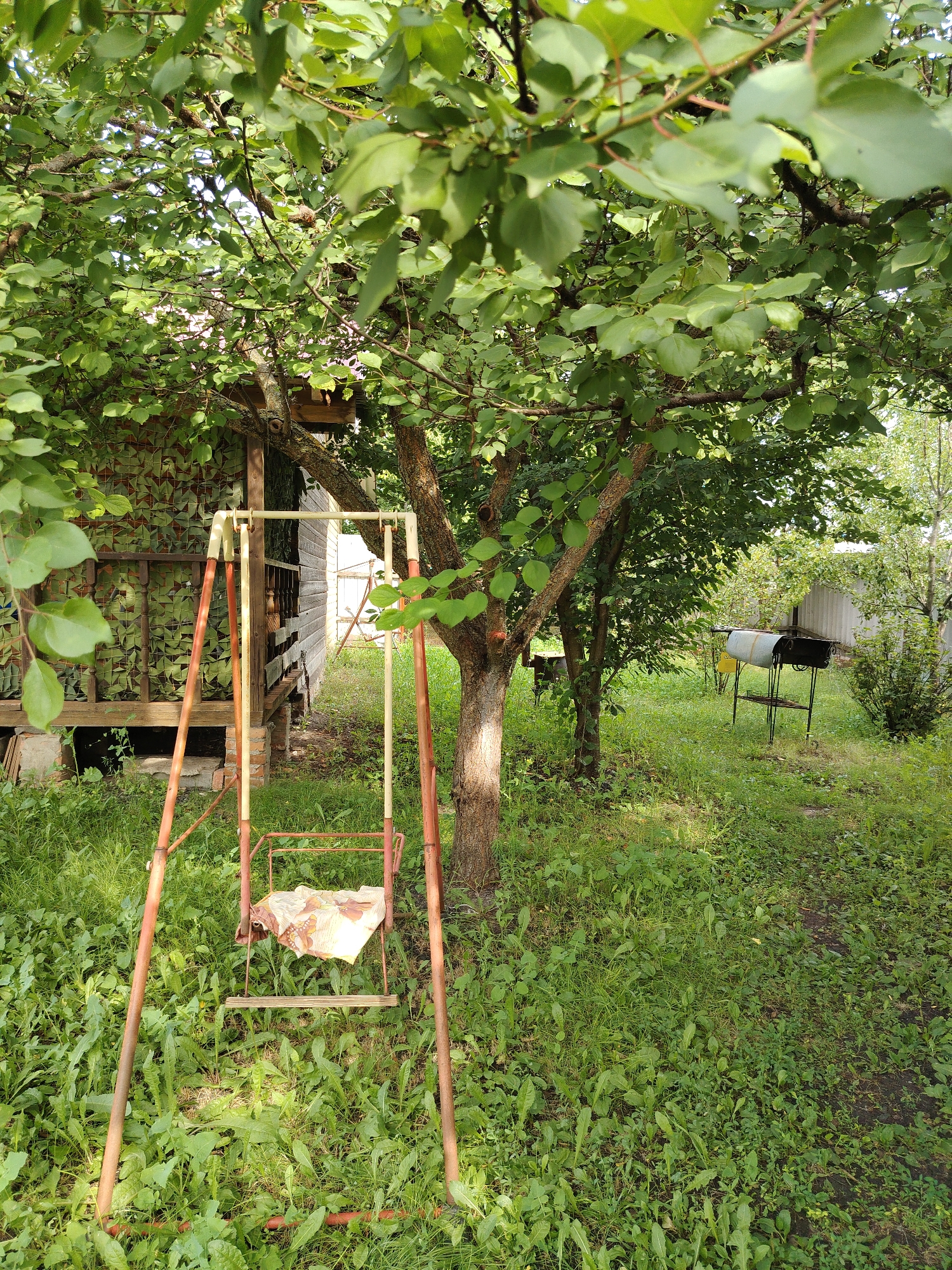 In Volokonovka, Russia, a tranquil backyard scene unfolds beneath the dappled shade of a mature tree. An old, rusty swing set stands prominently in the foreground, its weathered frame adorned with a faded, patterned fabric. To the left, a wooden structure, partially covered by camouflage netting, suggests a rustic shed or outbuilding. Beyond this, the scene opens to a verdant lawn interspersed with various plants and grasses, leading to a barbecue grill with a metallic body and a black grate. In the background, a simple fence delineates the property, with more trees and foliage creating a sense of natural enclosure. The overall impression is one of a quiet, lived-in outdoor space, typical of a Russian dacha or countryside home.