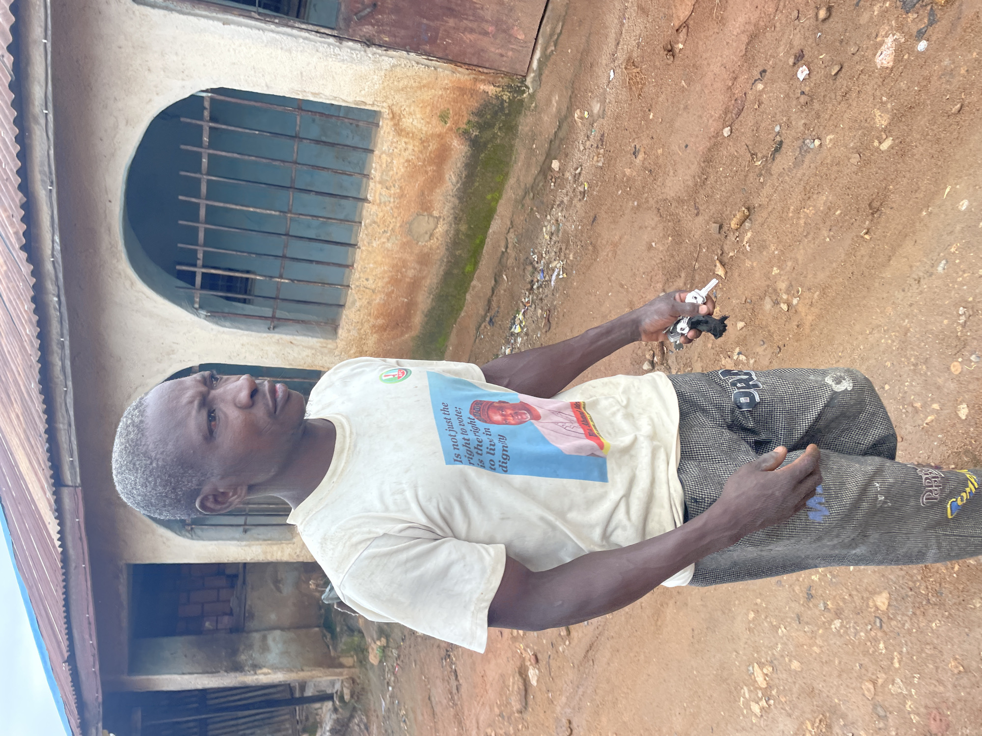 A medium shot shows a man standing on a dirt road in front of a weathered building with a corrugated metal roof. He has short, light-colored hair and is wearing a white t-shirt with a political slogan and a picture of a man in traditional attire. The t-shirt has a small circular logo in the upper right corner. The man is holding a set of keys in his right hand. The building appears to be a residential structure, with arched windows covered by metal bars. The ground is uneven and scattered with debris. The overall scene suggests an outdoor environment in a developing area.