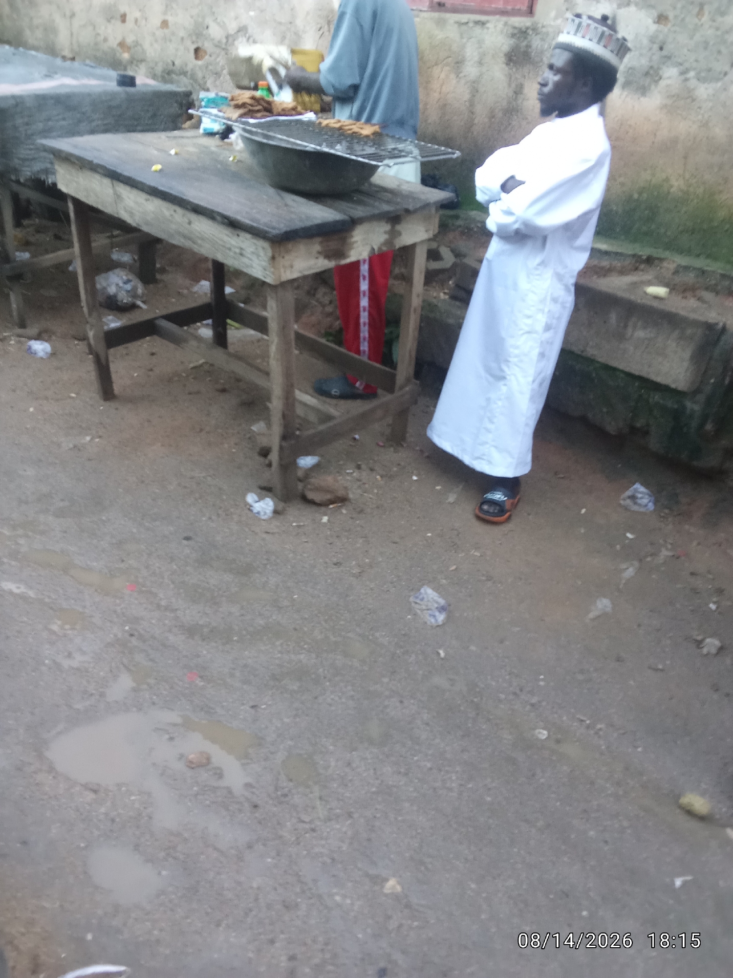 This is a photograph taken on August 14, 2026, at 18:15, showing a street scene in Jos, Nigeria. In the foreground, the ground is a mix of asphalt and dirt, with several puddles and scattered debris. A man wearing a white robe and a patterned kufi hat stands with his arms crossed, facing left. Behind him, a wooden table holds a metal basin filled with food items, possibly grilled meat. Another person, partially visible, is wearing a light blue shirt and red trousers, also near the food stall. The background shows a textured, light-colored wall and some green foliage. The scene appears to be an outdoor food vending area.