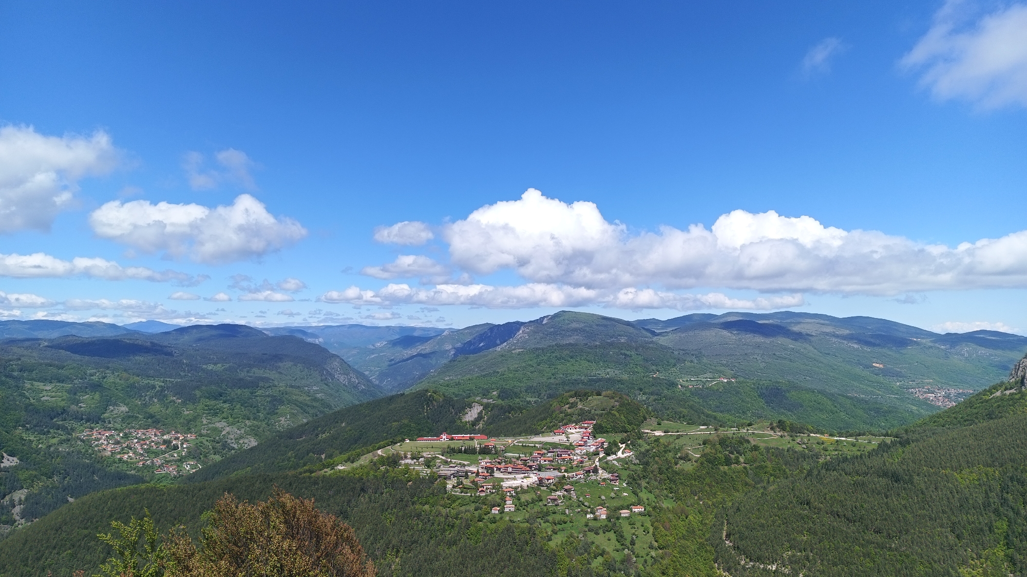 The image captures a panoramic view in Sini Vrah, Bulgaria. The scene is dominated by a series of forested mountains and valleys stretching into the distance under a blue sky with scattered white clouds. In the foreground, a settlement with red-roofed buildings is visible, nestled on a ridge. The landscape is mostly green, indicating a temperate climate. No people are visible in the shot. The composition suggests a viewpoint from an elevated position, possibly a mountain peak or high ground.