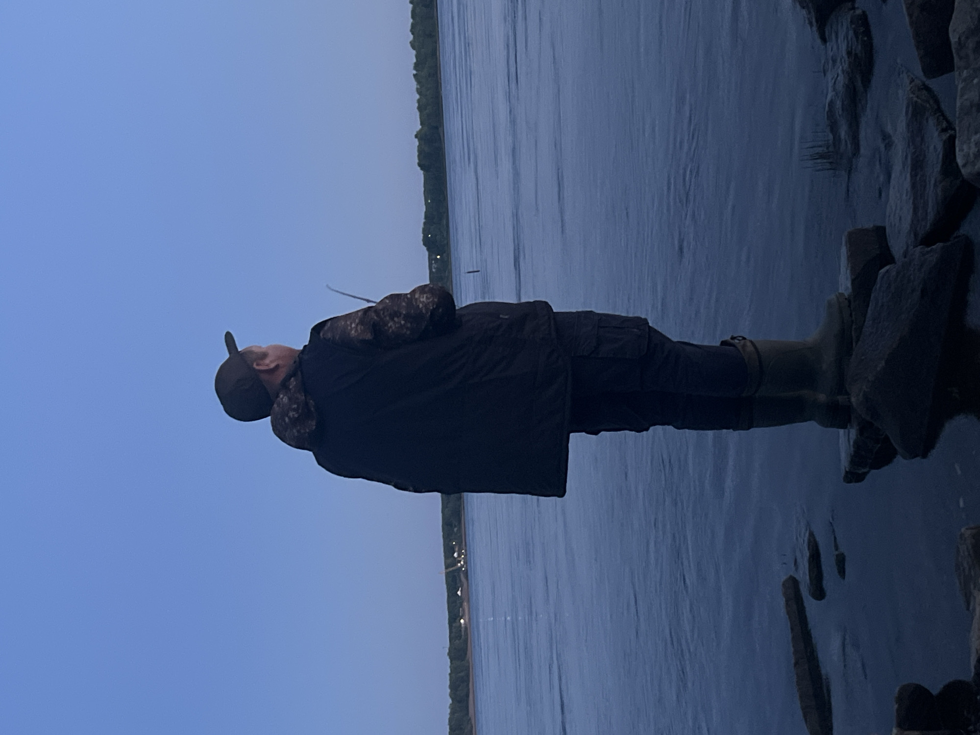 A man stands on rocks at the edge of a body of water, facing away from the camera. He is wearing a dark baseball cap, a dark jacket with camouflage accents on the shoulders, dark cargo pants, and tall rubber boots. He appears to be holding a fishing rod, with the tip of the rod visible extending into the frame. The water is calm, with ripples on its surface. In the distance, across the water, a treeline is visible, with several structures and lights suggesting a settlement on the far bank. The sky is a clear, pale blue, indicating it is either early morning or late evening. The overall lighting is dim. The location is the Kama River in Chaikovsky, Russia.