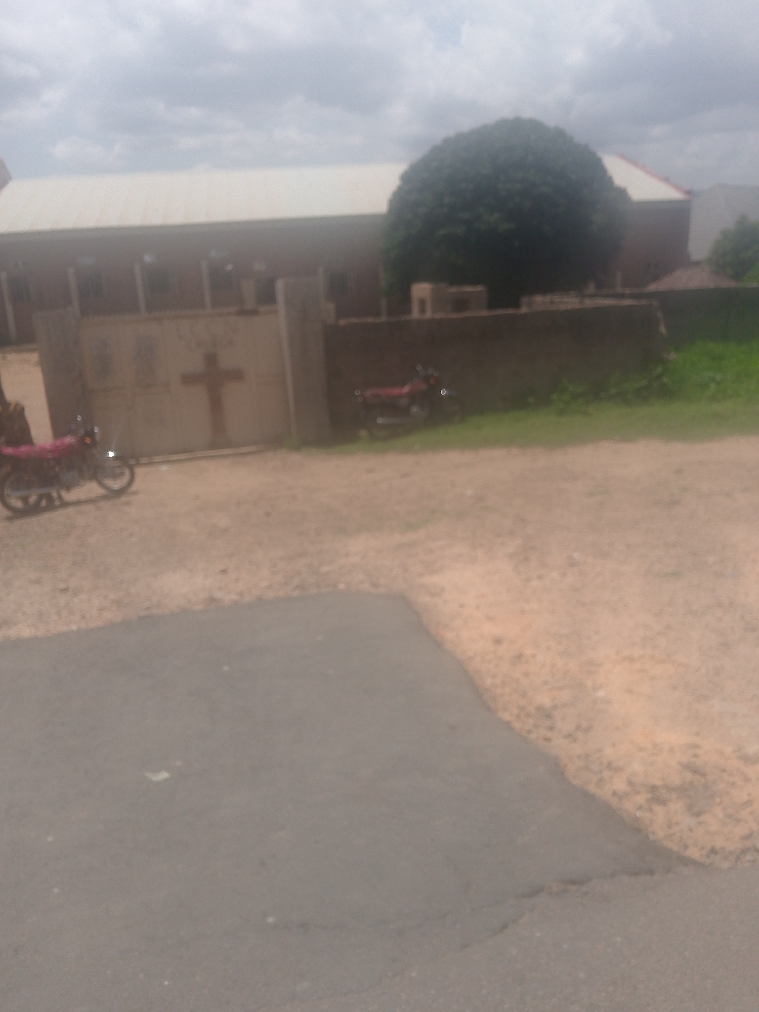 This image depicts an outdoor scene in Chum, Nigeria, under an overcast sky. In the background, a large building with reddish-brown walls and a light-colored, possibly corrugated metal, roof is visible. A prominent light-colored gate stands in front of this building, centrally featuring a large, dark-colored cross. Faint, blurry text, possibly "LCCM CHURCH" or similar, is discernible above the cross on the gate. To the right of the building, a large, dense, spherical green tree is prominent. A low, dark wall extends horizontally across the mid-ground, positioned in front of the building and the tree. Behind this wall, to the right, a patch of green grass is present.
The mid-ground is dominated by a large, unpaved expanse of light brown, dusty ground. On the far left, a red or maroon motorcycle is parked, facing towards the right, with a silver-colored engine or exhaust pipe visible. A partial human figure, specifically a lower body and leg, is faintly visible to the left of this motorcycle. A second red or maroon motorcycle is partially visible further back on the right, positioned behind the low wall. The immediate foreground consists of a dark grey paved road, which curves from the left edge of the frame towards the bottom right, transitioning into the dusty, unpaved ground of the mid-ground.
