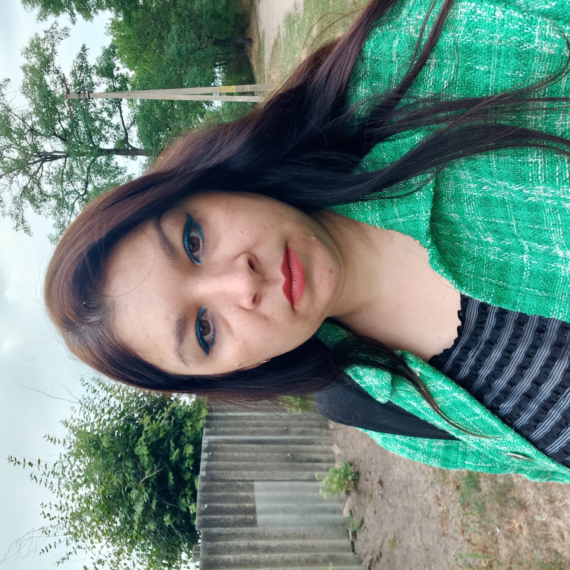 The image is a close-up, outdoor, selfie-style photograph of a woman with medium-length dark brown hair, wearing a green textured jacket over a black and gray striped top. She has blue eyeliner and reddish-orange lipstick. The background consists of a grey corrugated fence on the left, dry grass and dirt on the ground, and trees and a utility pole with signage on the right. The overall environment appears to be a rural or suburban area, consistent with the city of Klyntsi, Ukraine. The sky is overcast.