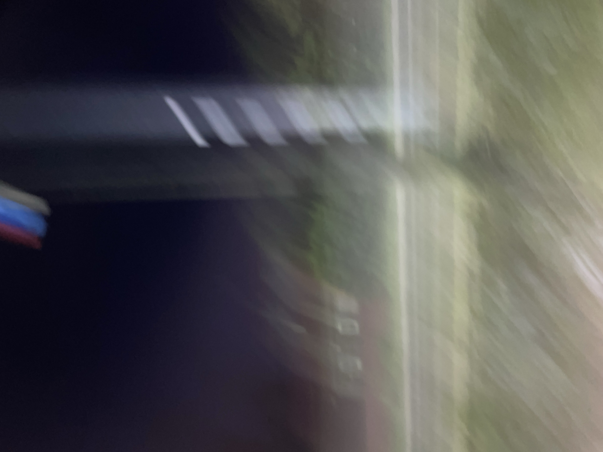 The image depicts a blurry night scene, likely taken from a moving vehicle. A tall, dark pole with white diagonal markings is visible on the right side of the frame, extending upwards into the dark sky. At the very top of the pole, faint red and blue elements suggest some kind of signage or lighting. To the left of the pole, a row of indistinct, blurry objects are visible, possibly streetlights or signs. A horizontal line, likely a road, runs across the middle ground, with a strip of green, blurry vegetation to its left. The overall impression is one of motion and low light conditions. The location is identified as Красные Баки, Russia, implying this is an outdoor scene within the city.