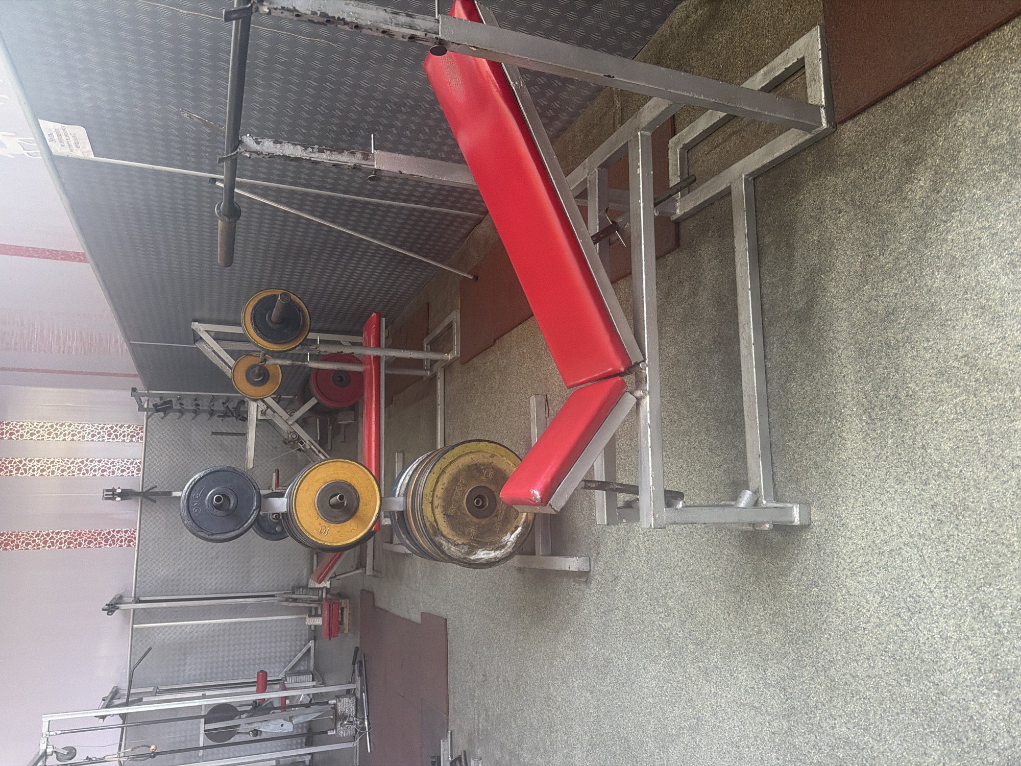 The image captures an indoor gym or weightlifting area, devoid of people or ongoing activity. The primary subjects are various pieces of gym equipment.
In the foreground and center, a robust, silver-painted metal frame supports a bright red upholstered weightlifting bench, which appears to be adjustable with an inclined backrest. The metal structure shows some signs of use.
To the left of the bench, an assortment of barbell weight plates is visible, stacked on holders. These plates come in different colors, including yellow, red, and black. Several yellow plates are marked with the number "10", and a black plate is clearly labeled "15 KG". Another yellow plate partially shows the text "COCOA...10". The weight plates display considerable wear, including rust and dirt, suggesting heavy use.
The floor is covered with a mottled gray or greenish-gray textured material, possibly carpet or specialized flooring. Dark red rubber mats are laid in areas beneath the weights. The walls exhibit different finishes; one section is light-colored with a repetitive red pattern, while another appears to be a rough, textured gray material. The ceiling features a distinctive metallic checker plate pattern.
Further into the background, additional metal racks, barbells, and parts of what seem to be a cable machine or other workout stations are discernible. The overall atmosphere suggests a functional, well-used, and somewhat industrial-style workout space. A small, unreadable white sign is attached to the metallic ceiling. The time of day is not discernible, and no specific location cues other than the provided context of Shumen, Bulgaria, are visible within the image.