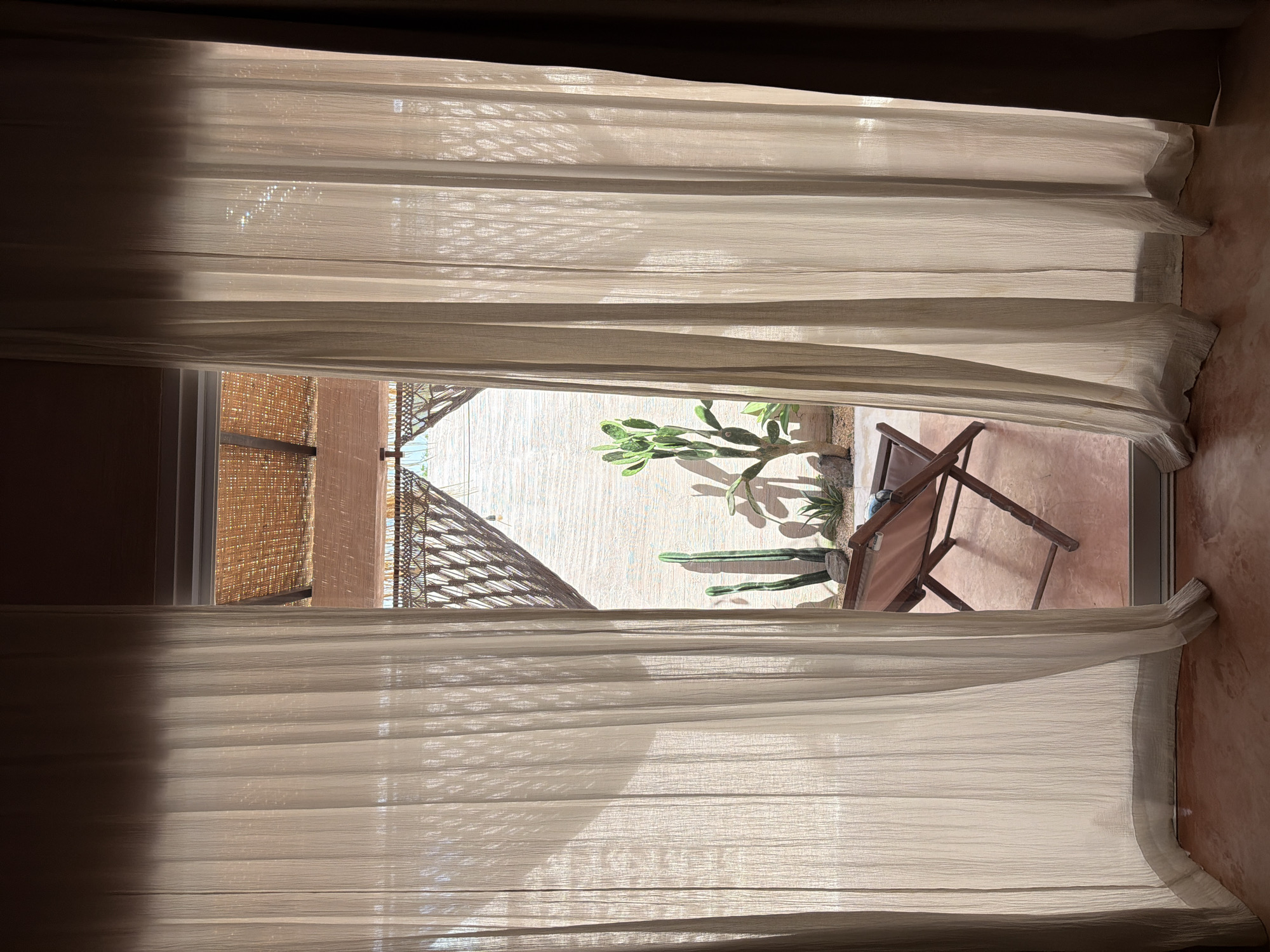 A partially open window reveals an outdoor patio scene. The window is framed by sheer, light-colored curtains that are partially drawn, allowing diffused sunlight to illuminate the interior. The patio features a light-colored wall, a cactus plant, a straw shade or roof overhang, and a brown wooden folding chair positioned on a light-brown tiled surface. The scene conveys an ambiance of casual indoor/outdoor living. The location is North Kuta, Indonesia.