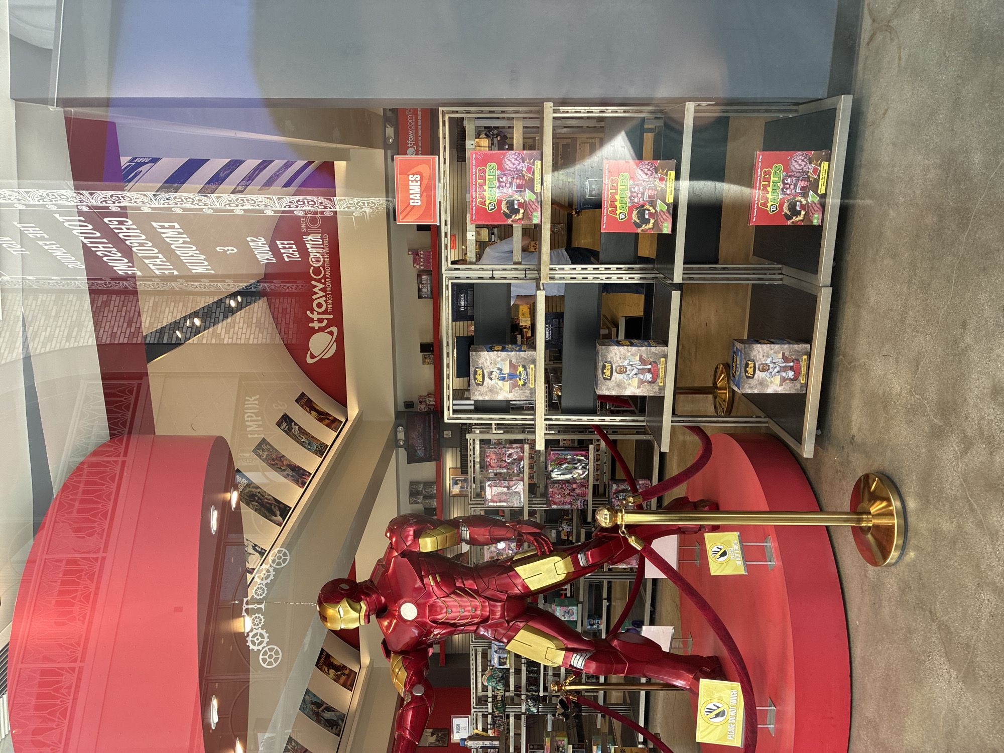 The image shows the interior of a comic book and collectibles store. A large, detailed replica of Iron Man stands prominently in the foreground, positioned on a red circular platform. The replica is made of shiny red and gold material, and is in a dynamic pose. Red velvet ropes and stanchions, with gold bases, surround the platform. Two yellow signs with black hand icons and the text "PLEASE DO NOT TOUCH" are visible on the platform.
Behind the Iron Man replica, shelves are filled with various merchandise. Some shelves display boxes of "Fallout" figures, and others hold boxes of "Apples to Apples" card games. The store appears well-lit, with overhead lighting visible. Reflections in the store window show some of the store's signage, including a red logo with "tfaw.com" and the tagline "THINGS FROM ANOTHER WORLD". There are also reflections of vertical stripes in blue and white on a pillar, and text that appears to be reversed.
In the background, more shelves of merchandise are visible, along with posters or displays on the walls. A glimpse of a person in a grey shirt is visible in the background between shelves, walking away from the camera. The floor is made of concrete. The overall impression is of a retail environment dedicated to pop culture and collectibles.