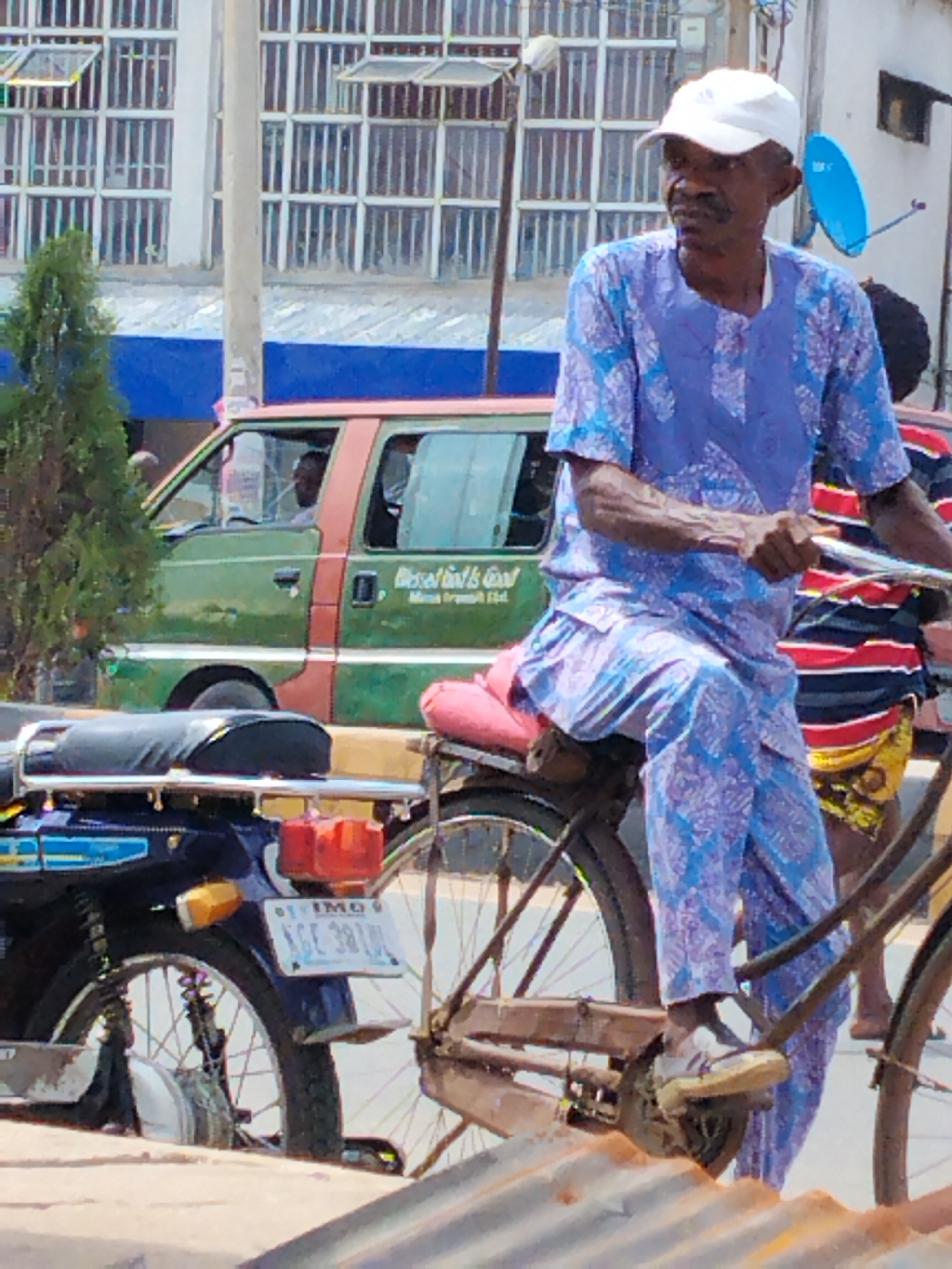 In Aba, Nigeria, an elderly man with a mustache, wearing a white cap and a blue floral-patterned top and pants, is seen riding a rusty bicycle. The man is pedaling the bicycle forward and is facing towards the right. A blue motorcycle with a license plate that reads "IMO NGE 381UL" is parked nearby. Behind the man is a green and red van, and a person wearing a red and black striped shirt is visible. In the background, there is a multi-story building with multiple windows. A blue satellite dish is mounted on one of the buildings in the background.
