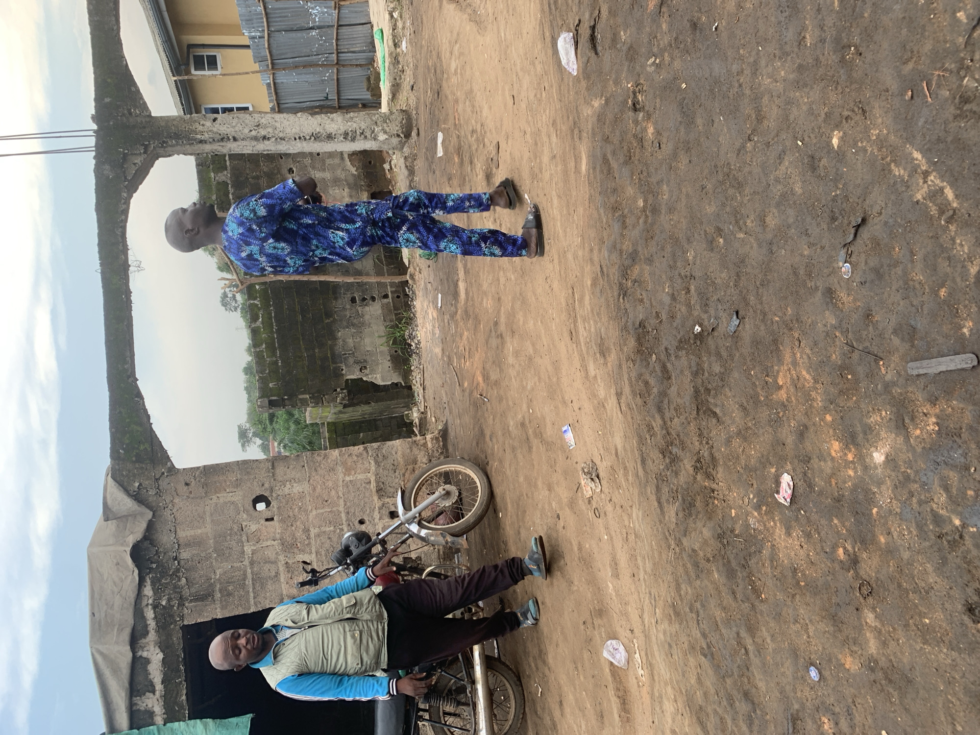 This is a medium shot, eye-level photograph taken outdoors in Shagamu, Nigeria. The scene depicts an unpaved, dusty area with several elements suggesting a possibly unfinished construction site or a maintenance yard.
In the foreground, the ground is a mixture of dark, packed dirt and lighter brown dust, with scattered debris including small pieces of paper, plastic, and wood.
Two men are present in the image. On the left, a man with a shaved head is standing next to a motorcycle. He is wearing a light blue zip-up jacket over a white shirt and dark trousers, along with dark sandals. He is looking downwards and slightly to his left. The motorcycle is dark-colored, with a visible exhaust pipe and spoked wheels.
To the right, a second man with a shaved head is standing and facing away from the camera, looking to the right. He is wearing a vibrant blue and teal patterned traditional African attire. He is also wearing sandals.
In the background, a dilapidated structure made of cinder blocks forms an archway. A weathered, light-colored tarp is draped over the top of the structure. Beyond this, another wall of cinder blocks is visible, and further back, a yellow building with windows and a corrugated metal fence can be seen. Trees and foliage are present in the distance, suggesting a semi-urban or peri-urban environment. The sky above is overcast with a few patches of lighter sky visible. The lighting is diffused due to the cloudy conditions.