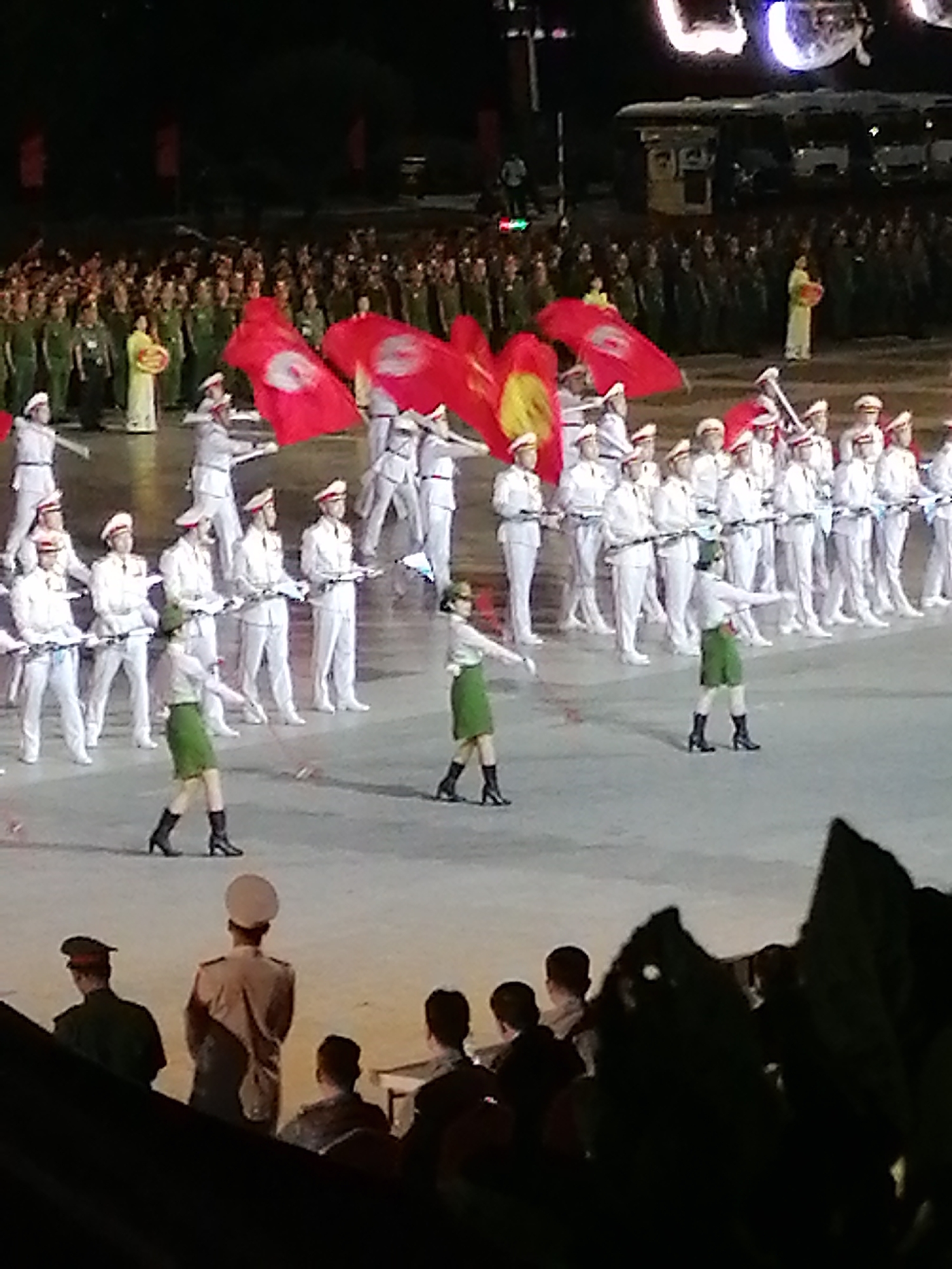 In Da Lat, Vietnam, at night, a large crowd gathers for a ceremony. In the foreground, four individuals, two of whom are clearly visible, are seated in silhouette behind dark foliage. Behind them, a group of people in uniform stand facing away from the camera. The central figure in this group wears a light-colored jacket and dark pants. In the middle ground, a formation of people in white uniforms, some holding rifles, stands in rows. Two individuals in the middle ground, wearing green skirts and black boots, are seen performing a dance or drill. Red flags with white circular emblems are held aloft by some participants in the middle ground. In the background, more uniformed individuals, likely spectators, fill the scene. Two figures in lighter colored clothing stand out among them. The environment appears to be an open, paved area, possibly a square or stadium, with a dark sky overhead.