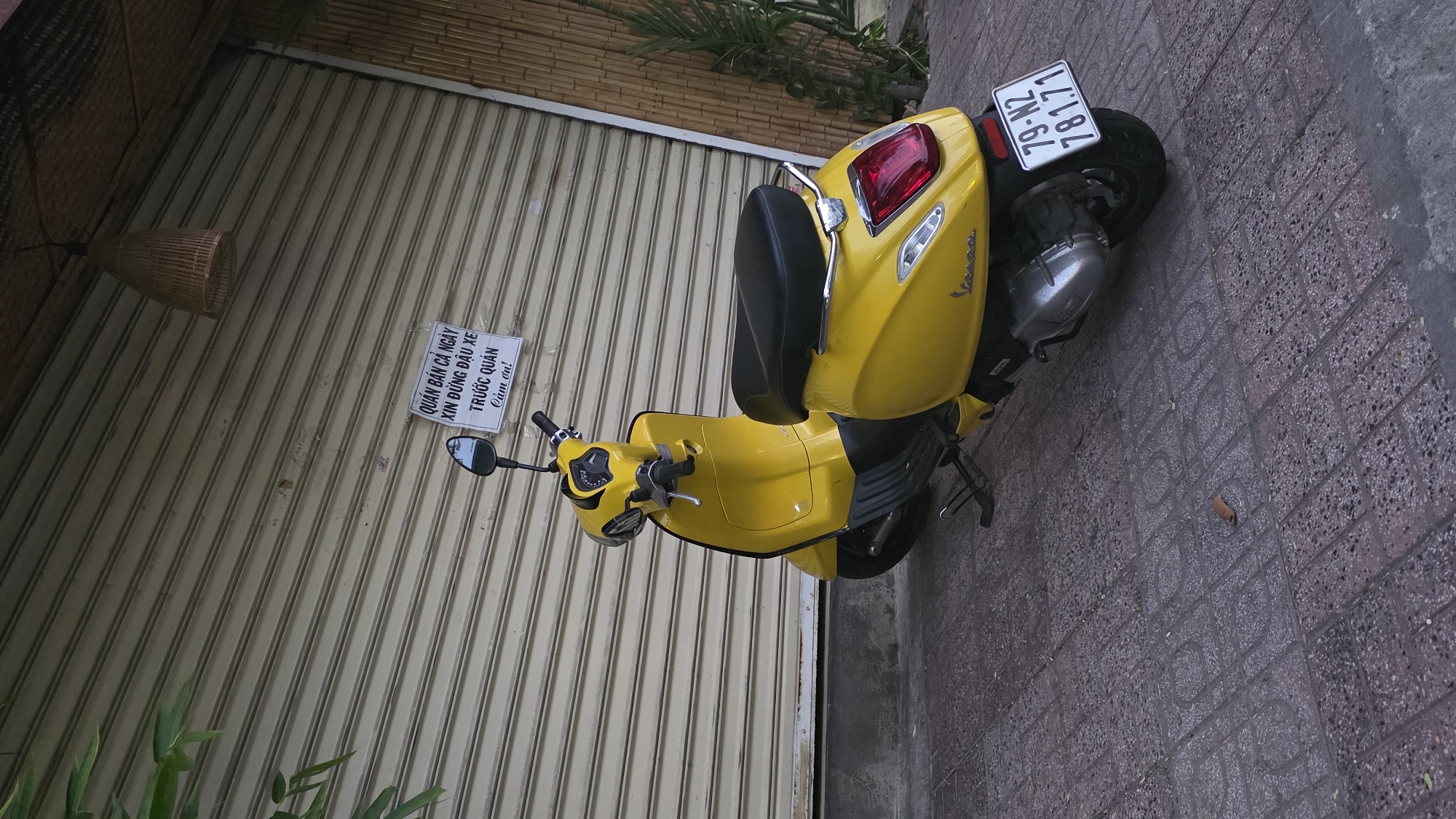 A yellow Vespa scooter is parked diagonally on a brick sidewalk next to a corrugated metal roll-up door, seemingly in front of a shop in Nha Trang, Vietnam. A small sign is taped to the door with Vietnamese text that translates to "Shop open all day. Please don't park in front. Thank you!". The license plate on the scooter reads "79-N2 781.71". A hanging woven basket lamp is visible above the scooter on the left side of the frame. Green foliage is visible along the top of the metal door.