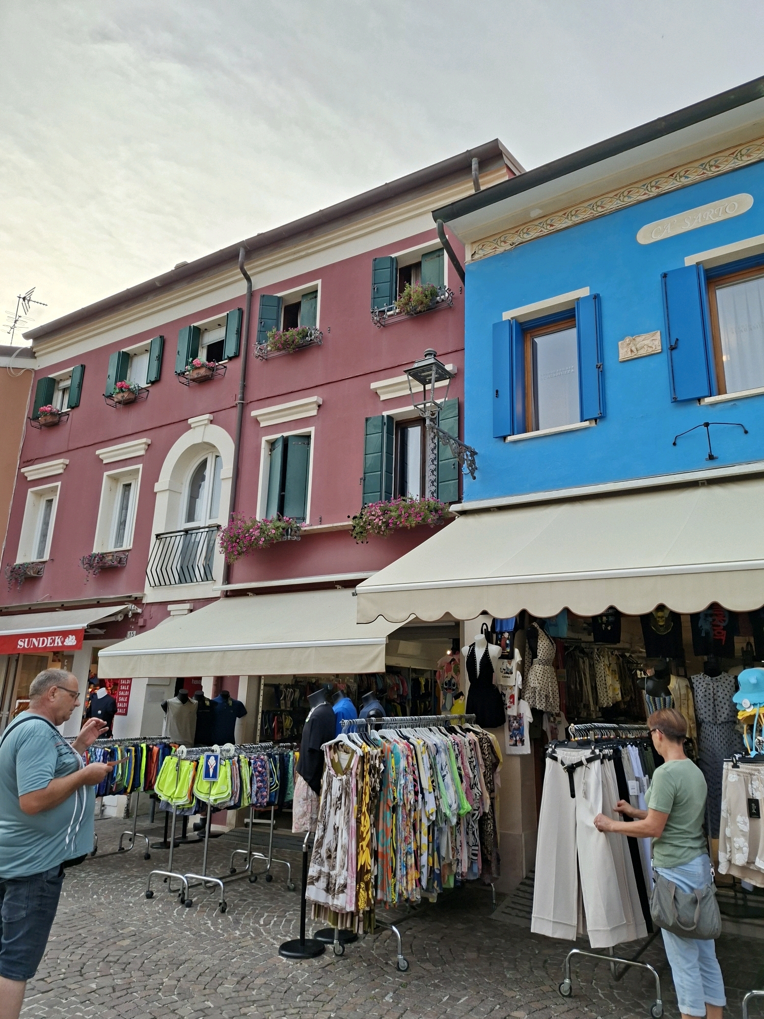 The image depicts a street scene in Caorle, Italy, with a focus on two adjacent buildings and a street-level market. The building on the left is painted a deep rose color with white trim, featuring several windows with dark green shutters, some adorned with flower boxes. The building on the right is a vibrant blue with blue shutters and decorative trim near the roofline. Above the shops, the building on the right displays the name "CA' SARTO".
In the foreground, a man wearing a blue t-shirt and dark shorts is looking at his phone, holding a small item in his other hand. He is standing in front of racks of colorful clothing, including shorts and patterned dresses. Further to the right, a woman wearing a green t-shirt and blue jeans is examining a rack of wide-legged white pants. Behind the people and clothing racks are store awnings and the storefronts themselves. The ground is paved with cobblestones. The sky is overcast.