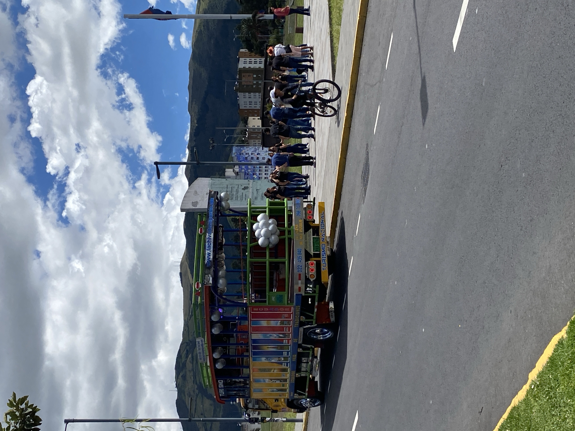 The image shows a colorful Chiva bus in Quito, Ecuador. The bus is parked on a city street next to a curb and a patch of grass. There are several people standing on the sidewalk next to the bus, including a person on a bicycle. In the background are some buildings, mountains, and a partly cloudy sky. A flag on a pole can also be seen in the background. The bus is adorned with lights, decorations, and balloons. The bus has the words "Turismo Nacional" and "Ecuador" on it.