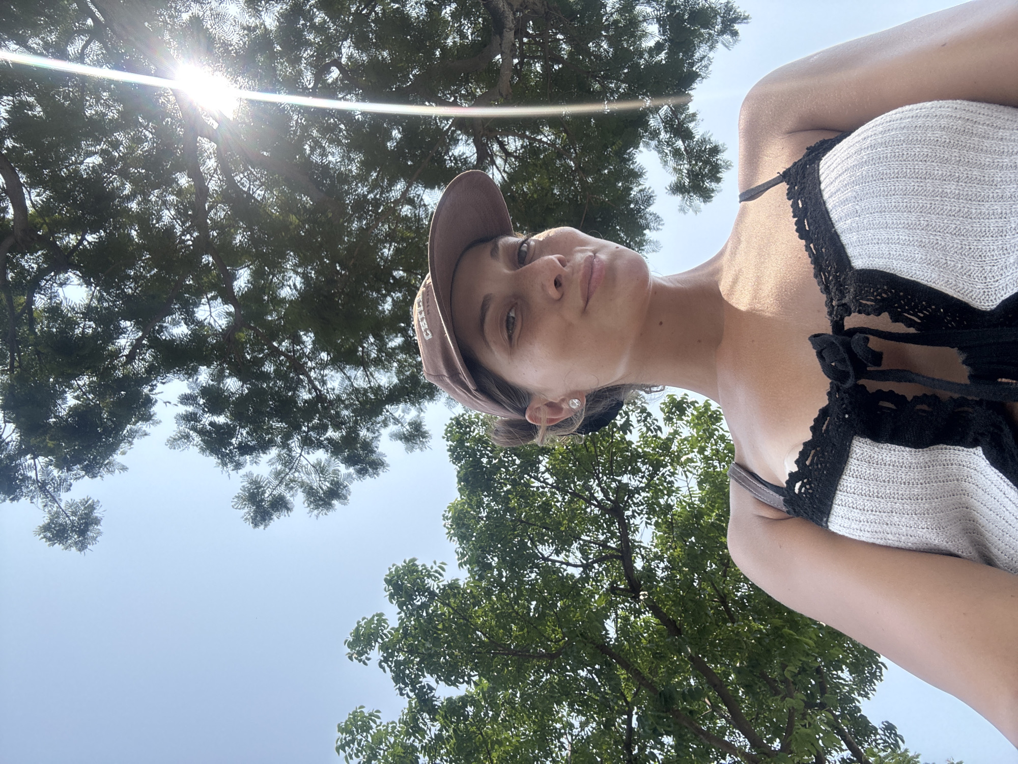 A woman wearing a brown baseball cap and a light-colored top with black lace trim is seen from a low angle. The sun is shining brightly, creating glare in the upper-left corner. Green trees and a blue sky fill the background. The location is potentially Đà Nẵng, Vietnam, based on geographical clues from the foliage.