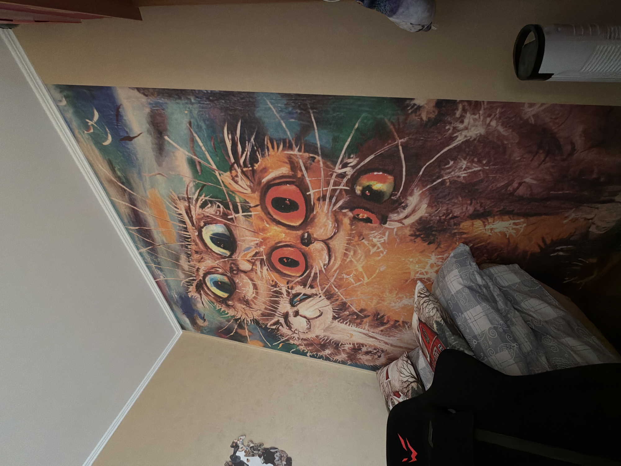 The image shows a bedroom interior. A wall mural featuring a surreal depiction of multiple cat faces dominates the scene. The mural's style is painterly, with visible brushstrokes and a mix of colors including orange, brown, blue, and green. A section of the room's wall and ceiling is visible, painted in neutral tones. In the lower portion of the frame, a gaming chair is present, with its back and seat in view. Several pillows with patterned fabrics are stacked near the chair. At the top of the image, there is a rolled-up item, potentially bedding or clothing, and a cylindrical object that could be a diffuser. The image was taken in the city of Shul'gino, Russia.