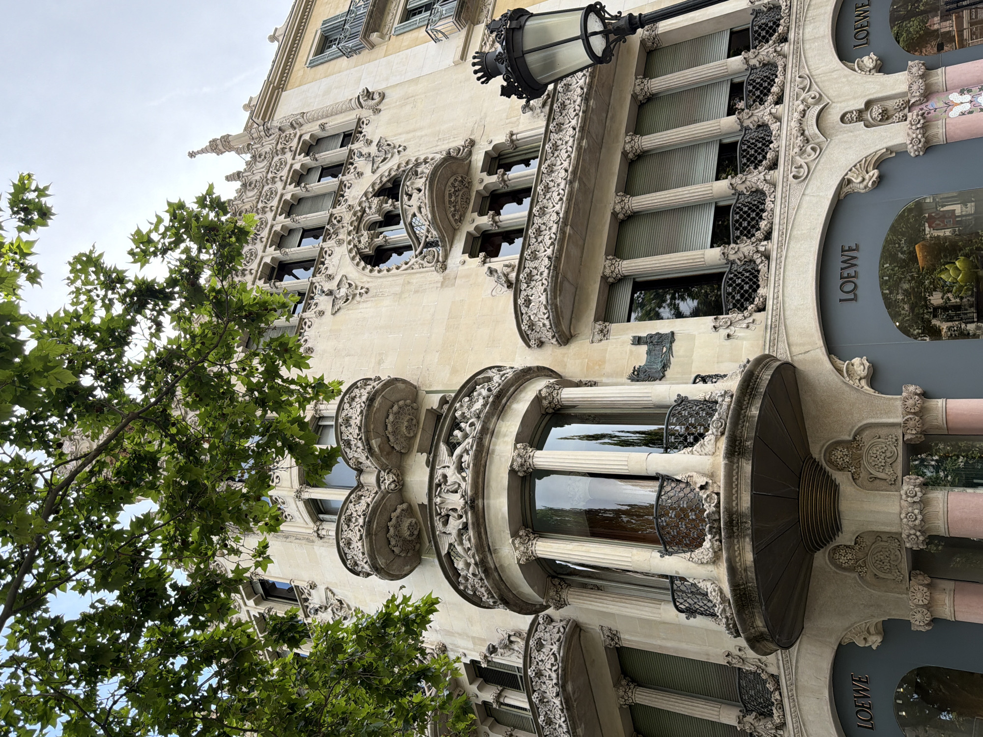 This is a low-angle outdoor shot of the exterior of a building in Barcelona, Spain, on a cloudy day. The building, located at Passeig de Gràcia, 92, is characterized by its ornate Art Nouveau architecture, featuring elaborate stone carvings, intricate balconies with decorative railings, and curved bay windows. The facade is made of light-colored stone, with some sections appearing weathered. Green leaves from a large tree partially obscure the upper portion of the building and branches are visible in the top left corner.
The ground level of the building houses a LOEWE store, with large, dark-grey display windows featuring the brand's name in gold lettering above them. The windows reflect the surroundings, showing greenery and the sky. In front of the building, on the right side of the image, stands a vintage black lamppost with a crown-shaped finial. The architecture displays classical elements like columns supporting the balconies.