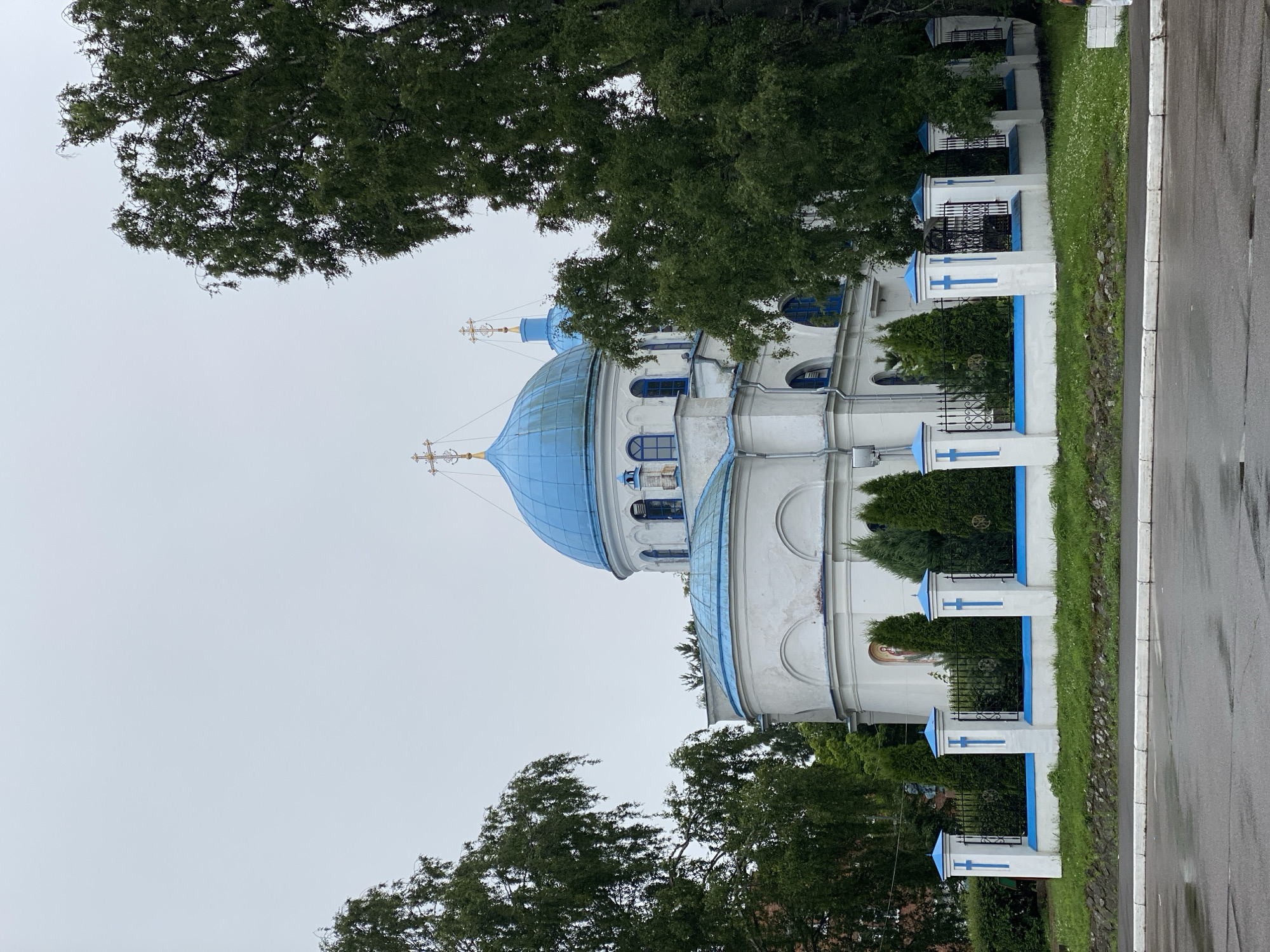The image shows a white church with blue domes and crosses, located in Priozersk, Russia. The church is surrounded by a white fence with blue crosses and a black metal gate. Green trees frame the church on both sides. In the foreground, a wet asphalt road runs parallel to the sidewalk. The sky is overcast and gray. There are no people visible in the image.