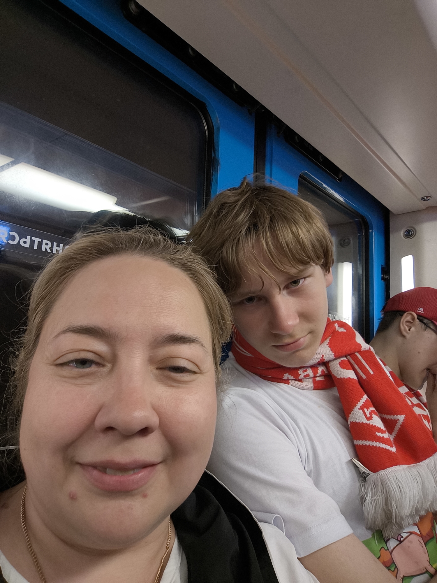 Three people are in the picture, two are clearly visible and the third is in the background. A woman is in the foreground, her face close to the camera. She has fair skin, blonde hair, and is wearing a gold necklace and a white shirt. Behind her is a younger person with blonde hair and a white shirt, wearing a red and white scarf. In the background, a person with a red baseball cap is visible. They are likely on a train or bus, as there is a window with reflection and blue interior trim visible. The location appears to be a city somewhere.