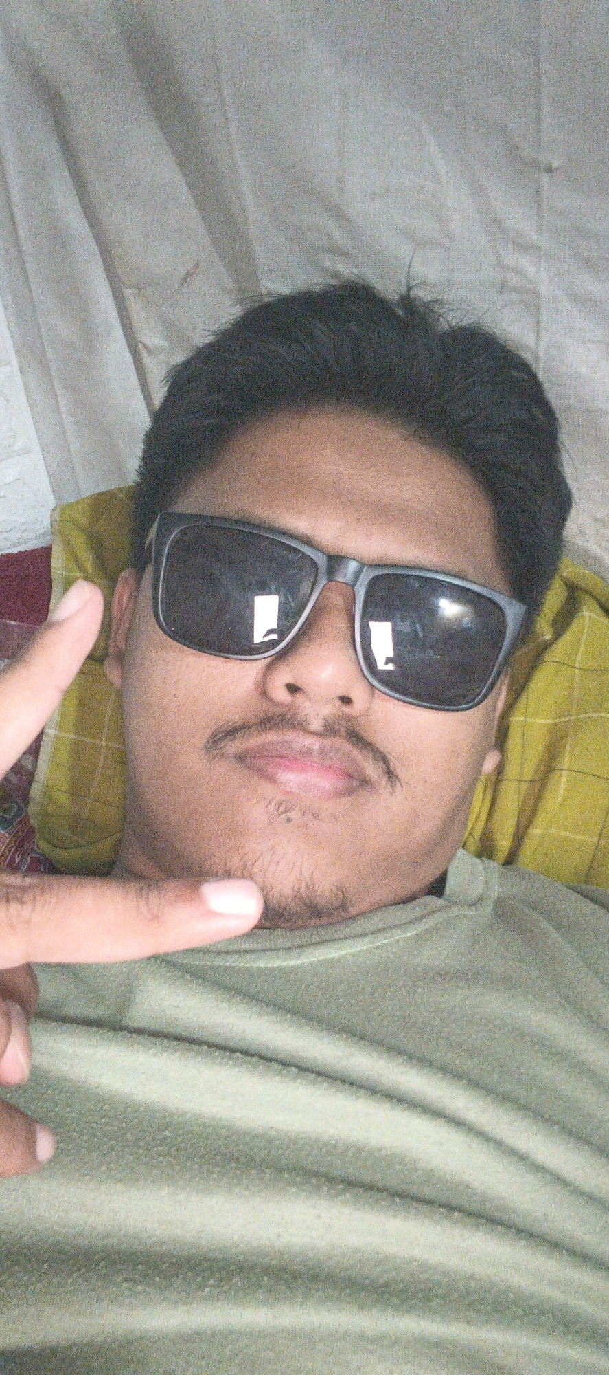 A selfie of a person is taken from a high angle, looking down at the camera. The person is lying down on a yellow pillow with a white and gray patterned blanket behind them. The person is wearing a green t-shirt and black sunglasses. Their hair is dark and short. They have a mustache and a small beard. The person is holding up their left hand with two fingers extended in a peace sign. The background is a white cloth with some folds and creases. The image is slightly grainy and the lighting is dim. It is not possible to determine the location in the city of Kemuning, Indonesia.