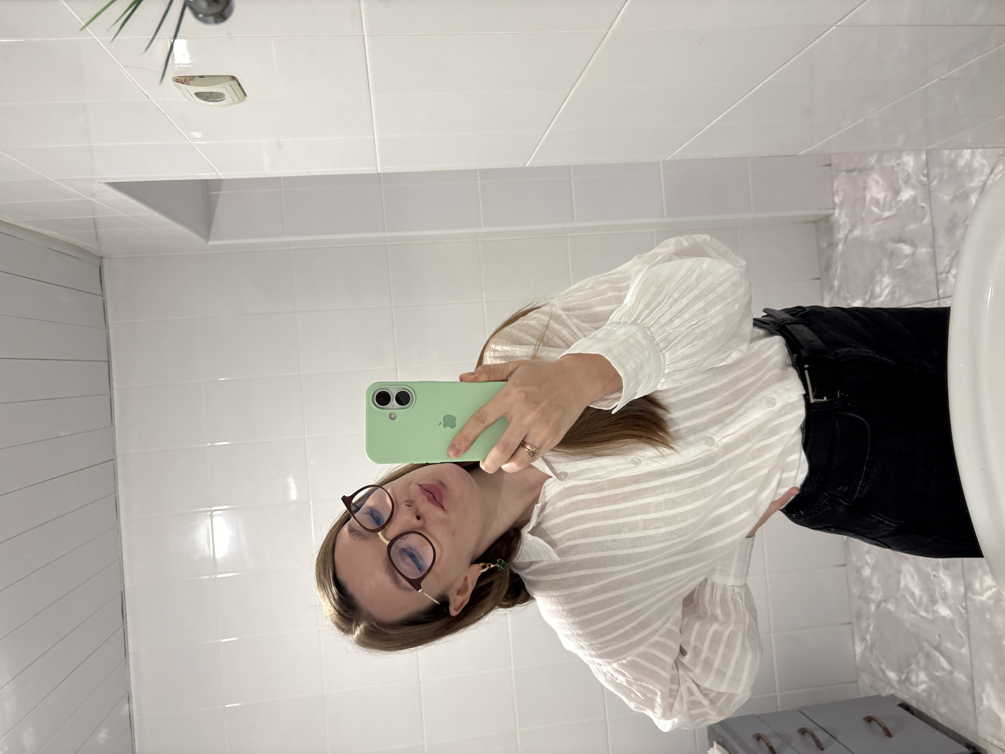 The image shows a young woman with blonde hair and glasses taking a mirror selfie in a bathroom. She is wearing a white striped blouse and black pants. She is holding a green iPhone in her right hand. The bathroom has white tiled walls and a tiled floor. There is a gray dresser to the left of the woman. A plant is visible on the right side of the image. The location is the city of Izhevsk, Russia.