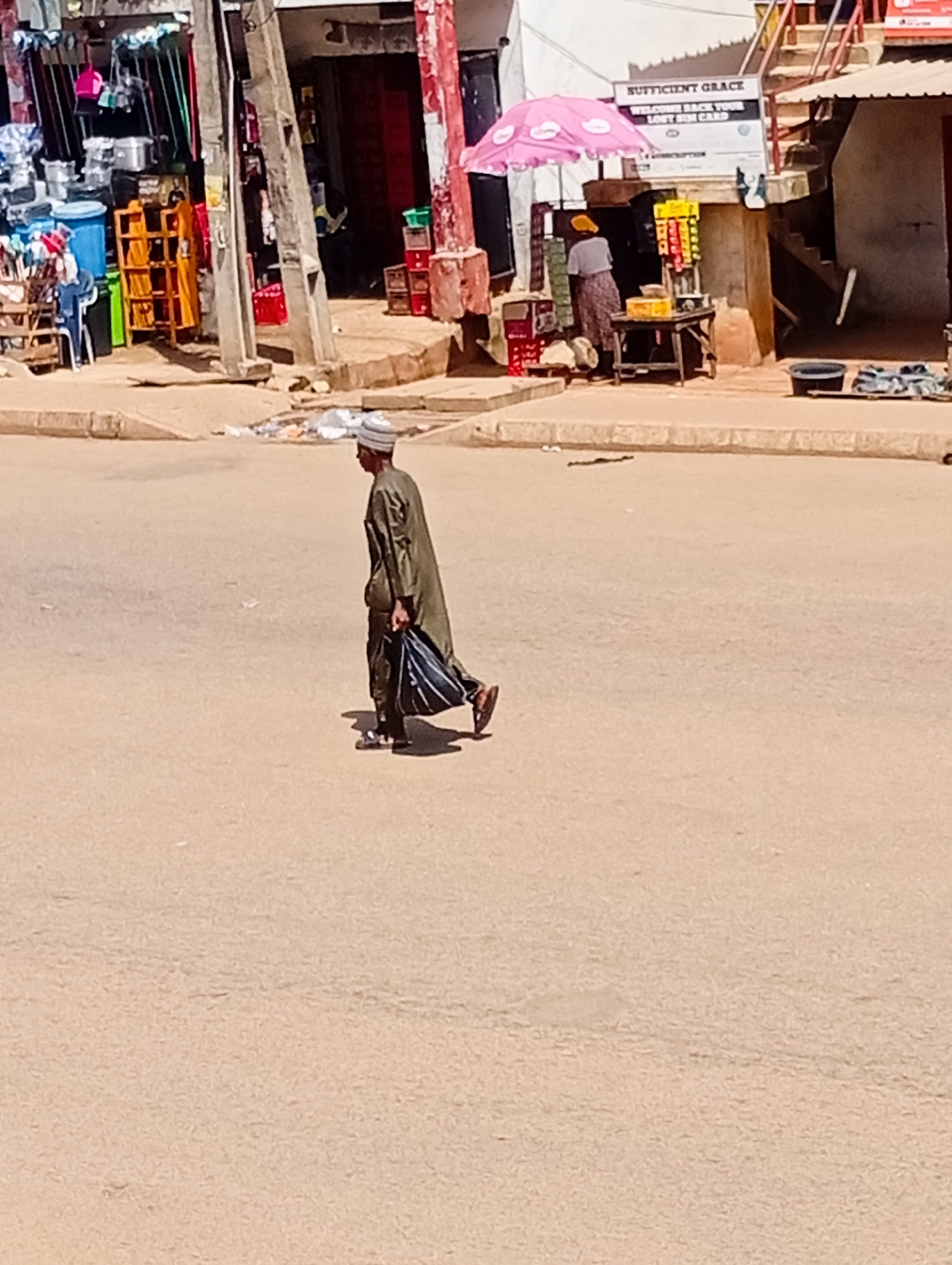A man wearing a green traditional robe and a white cap walks across a dusty street in Bukuru, Nigeria. He is carrying a striped bag in his right hand. The street is lined with shops and market stalls. In the background, a woman is standing under a pink umbrella, and there are various goods displayed for sale. The overall scene depicts a typical day in a Nigerian marketplace.