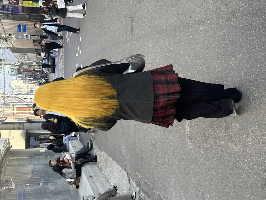 The image presents a detailed view of a bustling commercial street in the Hongdae district of Seoul, South Korea, observed during daylight hours. The primary subject is an individual, seen from the rear, walking forward on an asphalt street. This person has long, striking bright yellow hair, wears a dark charcoal-grey jacket, a red and black plaid pleated mini-skirt, and dark black trousers beneath the skirt. A silver-grey shoulder bag is carried on their left side.
On the left side of the street, a group of approximately five to seven people are seated on a low, grey concrete ledge fronting a building with large reflective glass windows, suggesting a retail or commercial space. One individual in the immediate left foreground is partially visible, also seated on this ledge, wearing light-colored trousers and black sneakers.
The background shows a densely populated urban environment with numerous pedestrians traversing the street in various directions. Multi-story commercial buildings line both sides of the thoroughfare. A vertical sign displaying Korean characters and the English text "LINE FRIENDS" is clearly visible on a building on the right side of the street in the mid-distance. Overhead utility wires are present across the street, and a few vehicles are partially discernible further down the road. The lighting indicates clear weather, likely during the afternoon.