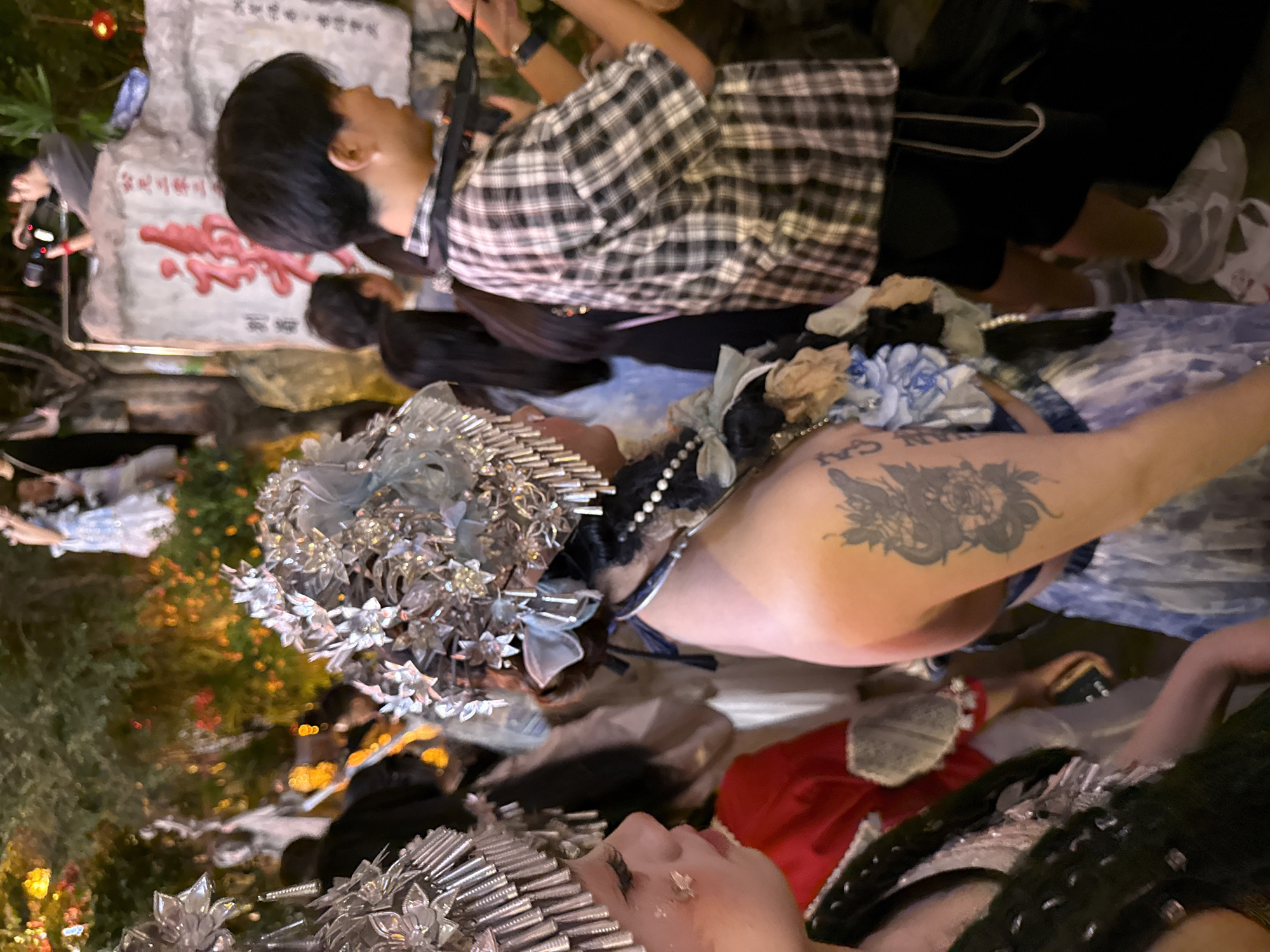 In Yuzhong District, China, the image captures a dimly lit outdoor scene, possibly during an evening event or festival. A young person in the foreground, on the left, is adorned with an elaborate silver headdress and white makeup, with faint glitter on their cheek. To the right, another person wears a similar, ornate headdress, featuring silver flowers and blue accents, and a beaded necklace. Their shoulder displays a tattoo of a serpent intertwined with a flower. This individual is dressed in a blue and white patterned dress, with a blue ribbon tied at the waist. In the mid-ground, a person wearing a black and white plaid shirt and dark shorts is visible, with a camera strap around their neck. They appear to be looking to the right. In the background, several other individuals are present, some with blurred features due to the low light and motion. Decorative lights, likely from lanterns or strings, create a warm, ambient glow, illuminating hints of foliage and a large rock with red calligraphy. The overall atmosphere suggests a cultural celebration or performance.