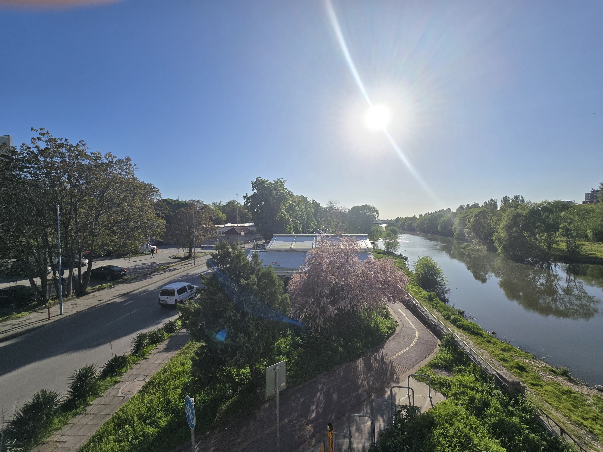 An elevated viewpoint captures a vibrant outdoor scene on a sunny day. A wide river or canal dominates the right side of the image, its calm surface reflecting the surrounding lush green trees that line its banks. A paved path, possibly for pedestrians or cyclists, runs along the near bank of the river, separated by a low concrete wall and metal railing.
On the left, a road parallels the river, featuring several parked cars and a white van moving away from the viewer. A lone person is visible walking on the sidewalk further up the road. The road is bordered by a grassy median with small plants and a sidewalk. A red fire hydrant stands near the sidewalk.
A prominent pink-flowering tree, possibly a cherry blossom, stands out near the riverbank, in front of a building with a white, translucent awning or roof. Text is visible on the awning, appearing to be "KASHMIR" (Кашмир) or similar, suggesting a restaurant or establishment. Abundant green foliage from various trees and shrubs fills the landscape, indicating springtime or summer.
The sky is a clear, bright blue, with the sun shining intensely in the upper right, creating a distinct lens flare and casting long shadows on the road and path, suggesting mid-morning or mid-afternoon. A blue circular sign with a white bicycle symbol is visible in the lower left foreground, indicating a bike path. Two small birds can be seen flying high in the distant sky on the right. The overall impression is one of pleasant, active outdoor urban life alongside nature.