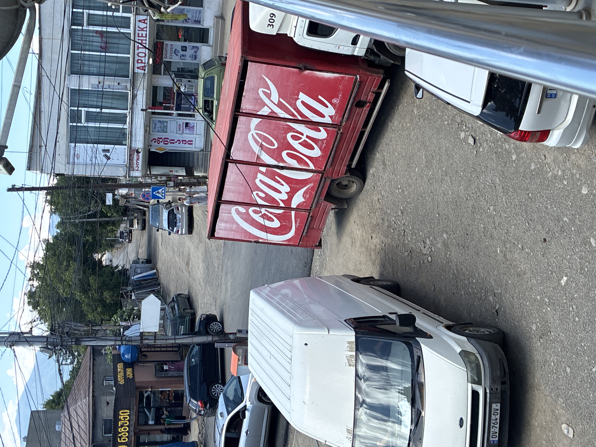 The image depicts a street scene in Khashuri, Georgia, with several vehicles and buildings visible. In the foreground, a white Ford Transit van is parked on a gravel road, its license plate reading "OV-764-OV". To the right of the van, a red Coca-Cola truck is parked, its side emblazoned with the iconic Coca-Cola logo. Adjacent to the truck, a white cab of another truck is visible, with the number "309" displayed on its side.
Further down the street, more vehicles are present, including a silver car and a green Lada hatchback. Buildings line the street, with one structure featuring a sign with "APOTEKA" written in large letters. Another building has signs with Georgian script. The street is unpaved and appears to be in a somewhat neglected state. Overhead, a complex network of electrical wires crisscrosses the sky. The overall impression is a bustling, everyday scene in a Georgian town.