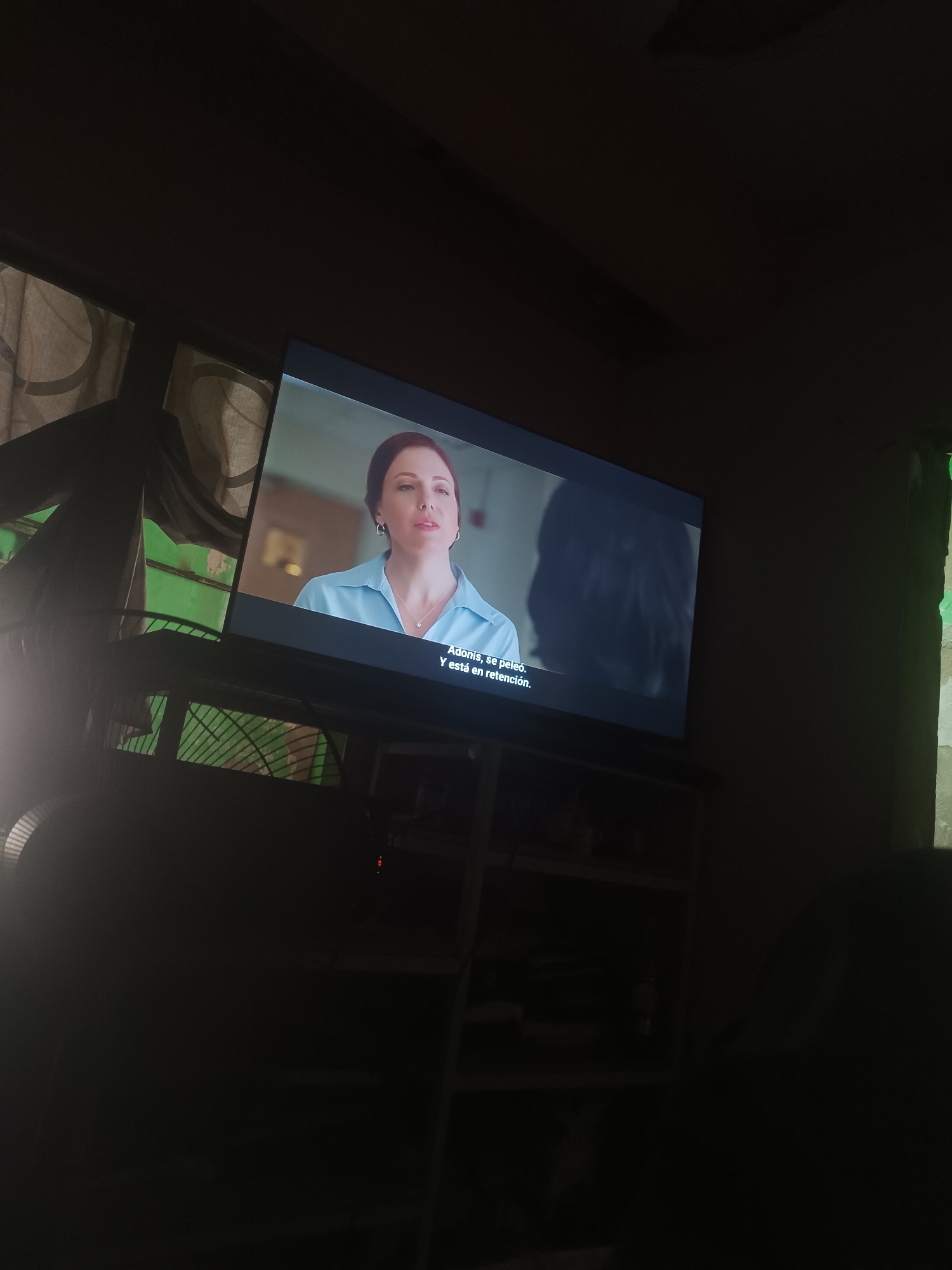 The image shows a television set in a dimly lit room. The TV is displaying a scene of a fair-skinned woman with reddish-brown hair wearing a light blue collared shirt and a necklace. She appears to be speaking. Subtitles in Spanish are visible at the bottom of the screen reading "Adonis, se peleó. Y está en retención." The TV is placed on a dark-colored stand or shelf. In the background, there are sheer curtains and what appears to be a window. There is a fan next to the window. The overall environment is dark and shadowy. The location within the city of Porlamar, Venezuela is not determinable from the image.