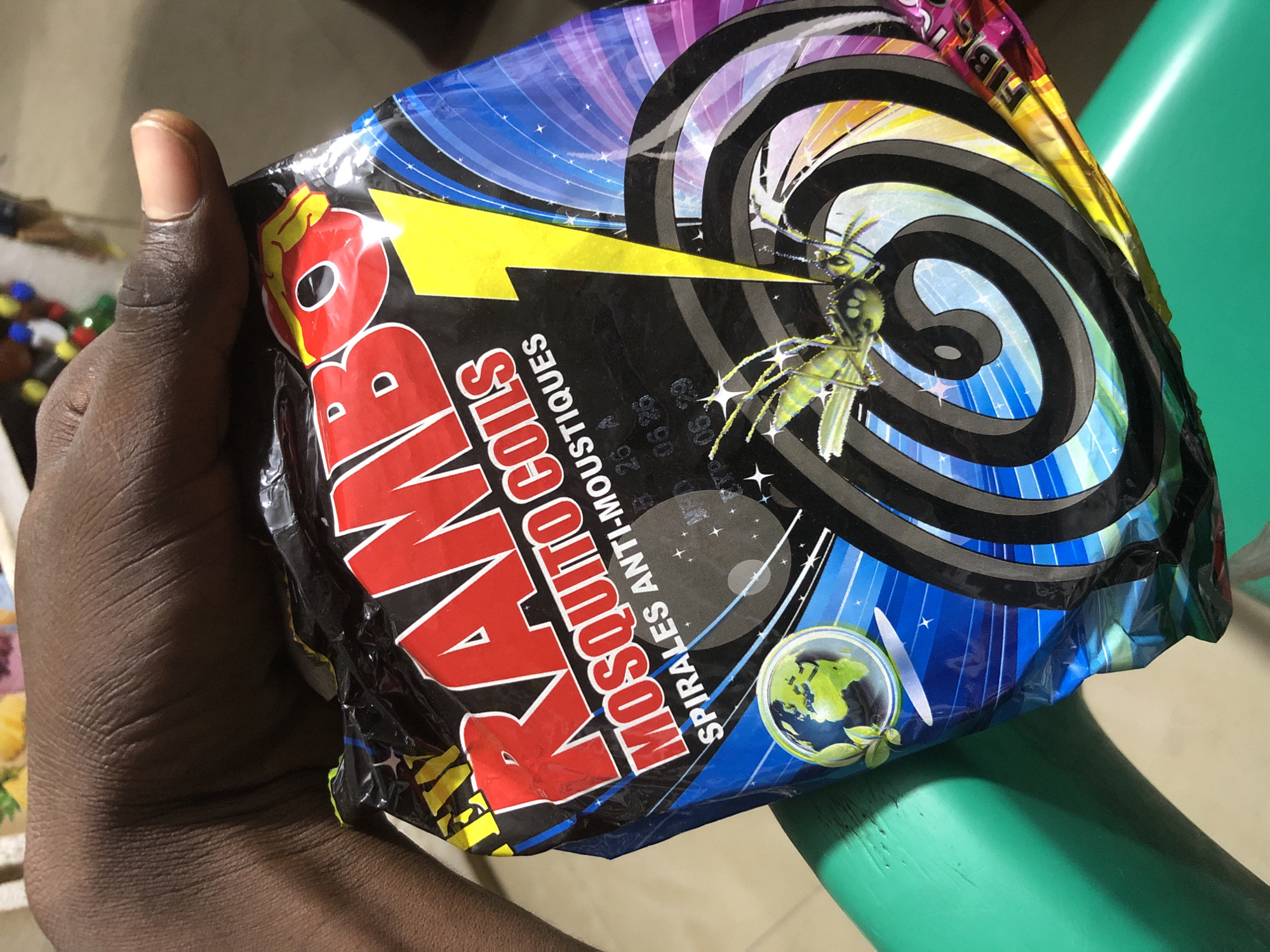 A dark-skinned hand holds a crumpled package of "RAMBO MOSQUITO COILS" which features a graphic of a lightning bolt and a wasp. The background is blurred, but a green object, possibly a chair, is visible. The image appears to be taken in a public space, possibly a market, in Ado, Nigeria.