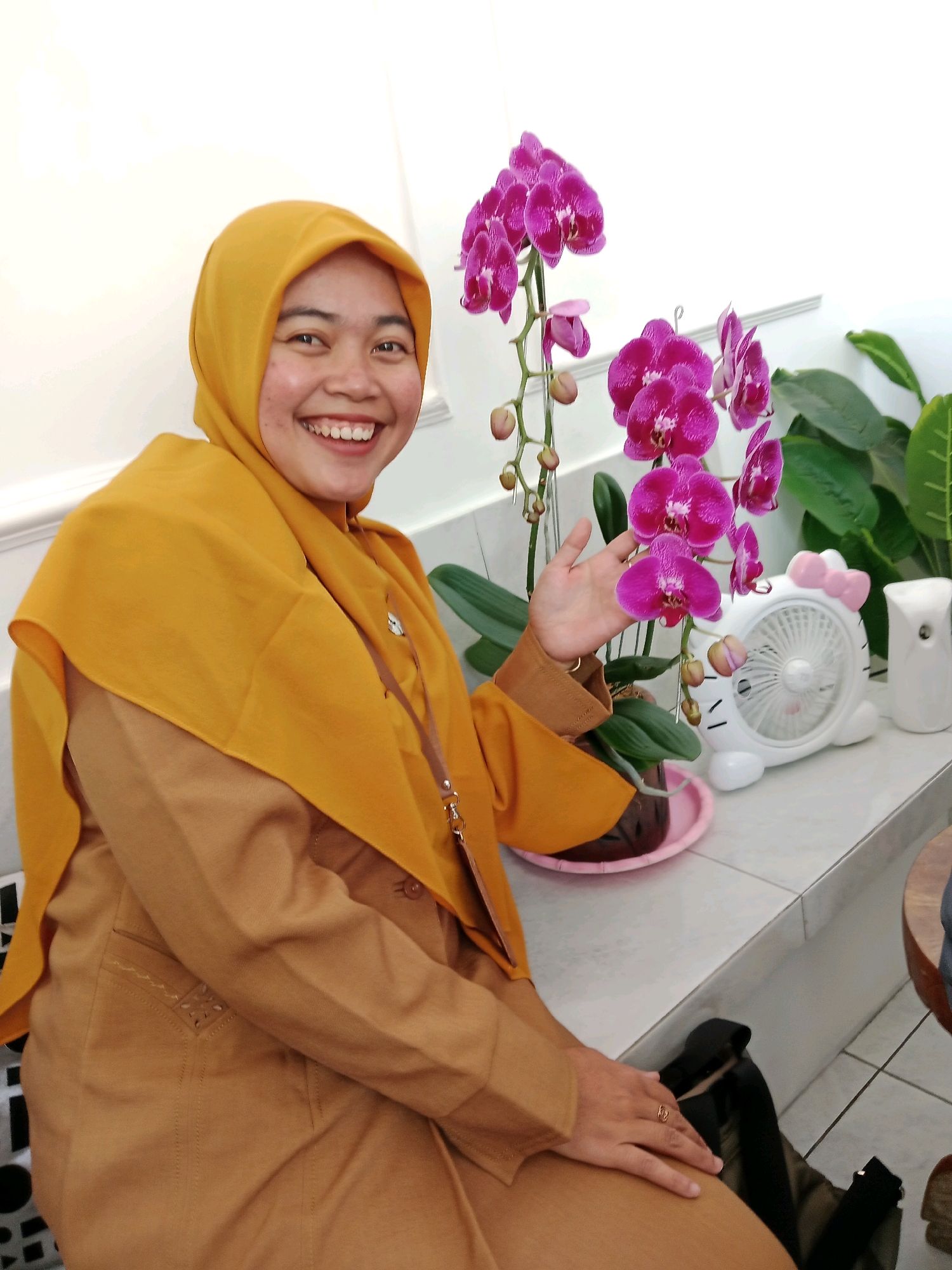A woman wearing a mustard yellow hijab and a tan uniform is smiling at the camera. She is sitting at a counter with pink orchids to her right. Behind the orchids is a white Hello Kitty-shaped fan and a white air freshener. The woman's right hand is resting on her lap, and a gold ring is visible on her right ring finger. The background is a white wall with a ledge running horizontally across it. The floor is tiled. The scene appears to be indoors, likely in Salatiga, Indonesia, given the context.
