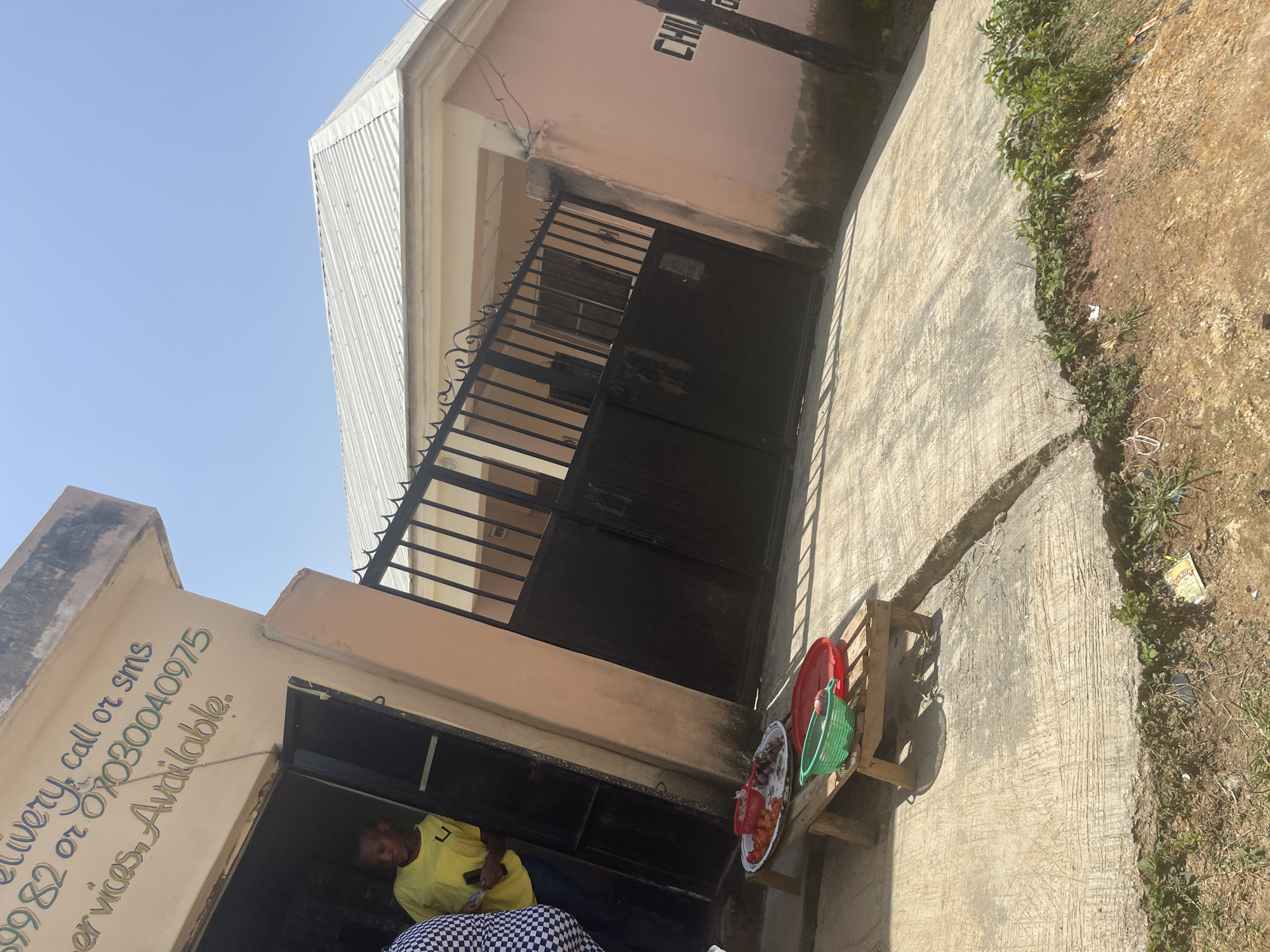 The image depicts a street view in Jos, Nigeria, under a clear blue sky. In the foreground, a small wooden bench holds a round tray filled with tomatoes and onions, alongside a green plastic basket containing an onion and a red lid. To the left, a person wearing a yellow t-shirt stands in the doorway of a building with beige-colored walls and a sign that reads "Delivery, call or sms 07030040975 Services, Available." The building's entrance is dark and partially obscured.
Behind the building, a light pink structure with a white metal roof is visible, partially enclosed by a black wrought-iron gate. The gate has decorative scrollwork at the top. On the wall of the pink building, partially visible text reads "CHILDREN." The ground in front of the buildings is paved concrete, with a section of uneven, cracked pavement in the mid-ground. A strip of dry earth with sparse green vegetation lines the edge of the paved area. The overall scene suggests a commercial or residential area with a small vendor display outside a shop or home.
